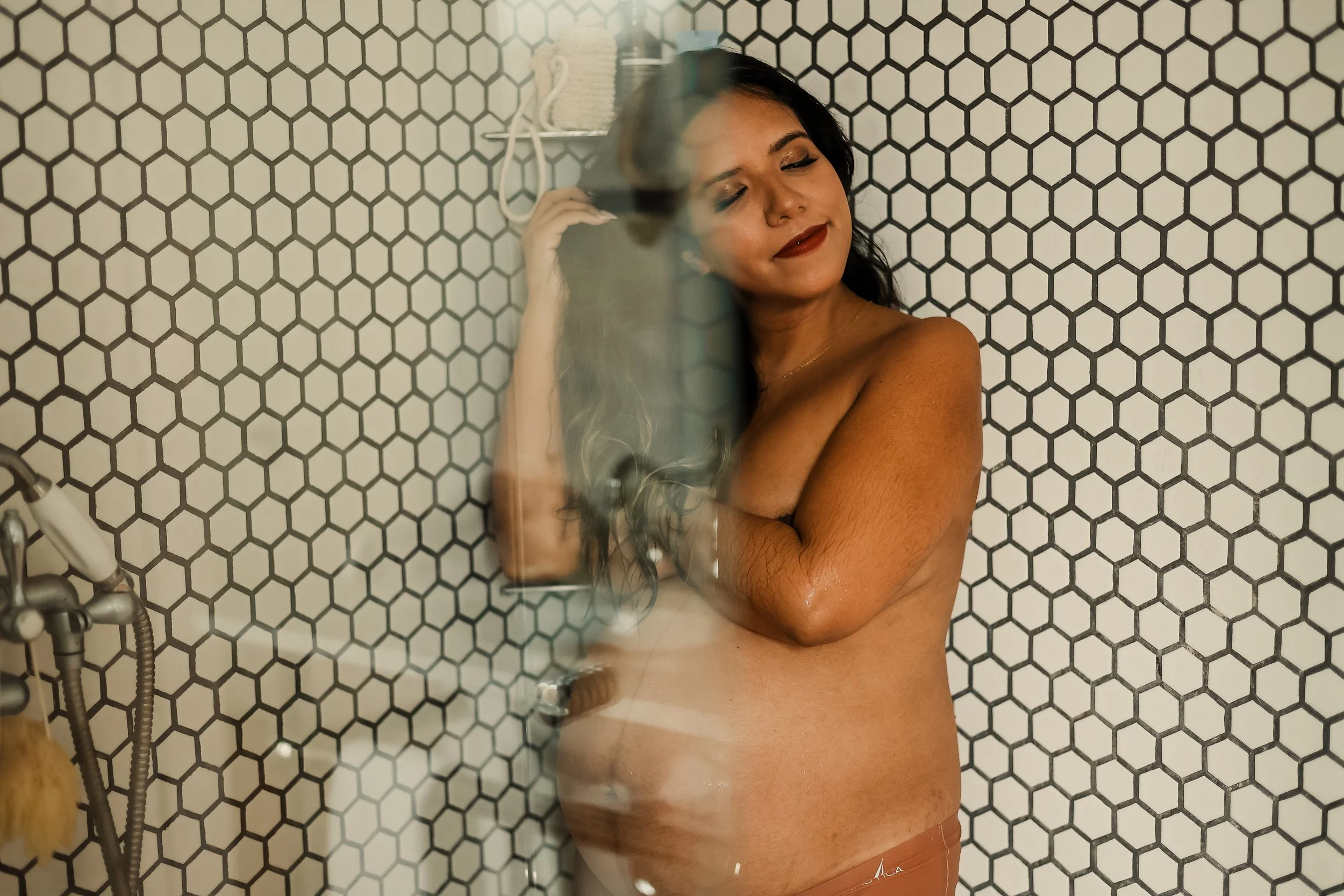 A woman with dark hair and makeup standing behind a glass shower door with a honeycomb tile wall in the background, smiling with her eyes closed.