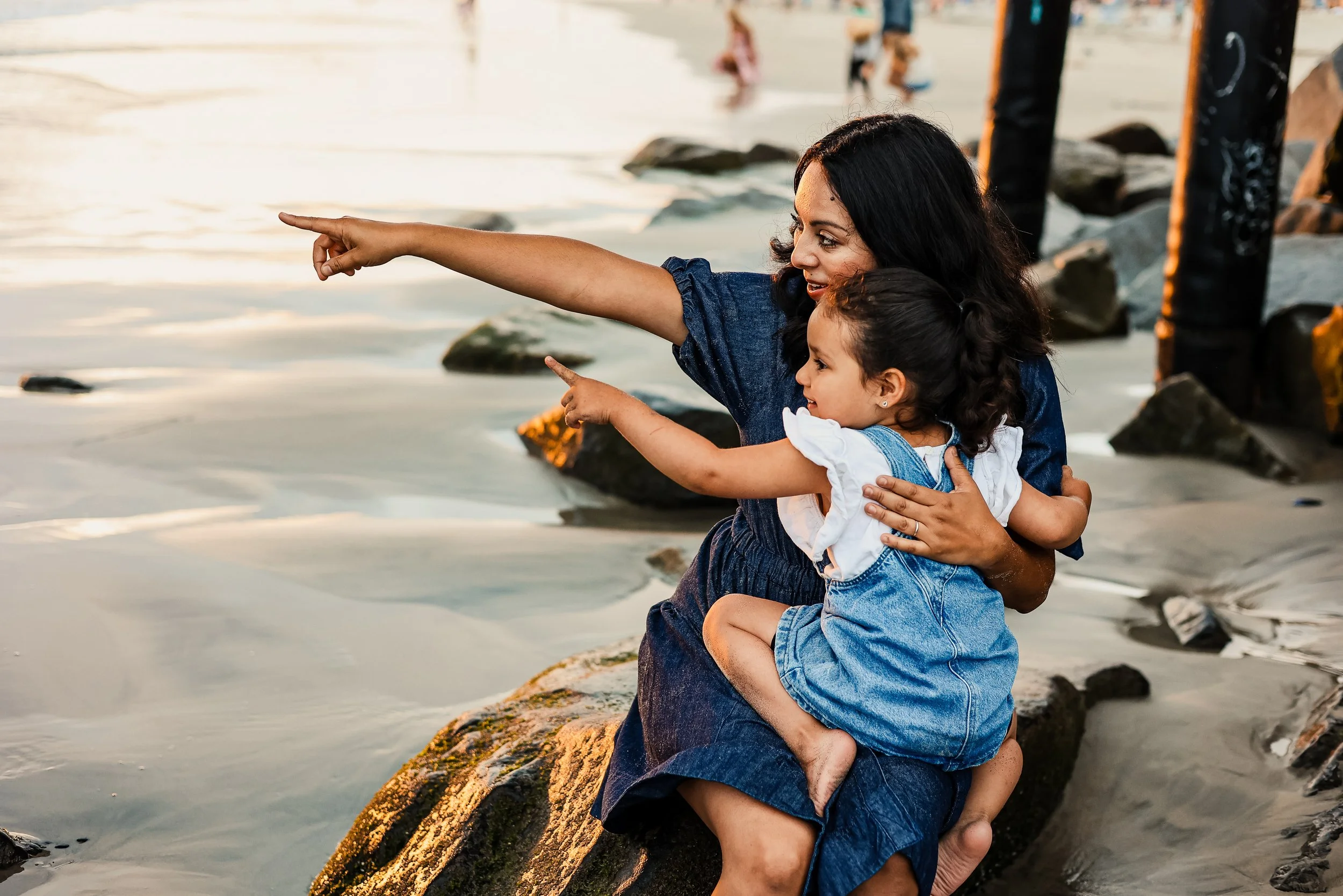 Mother and child sitting on rocks by the beach, pointing at something in the water, during sunset.