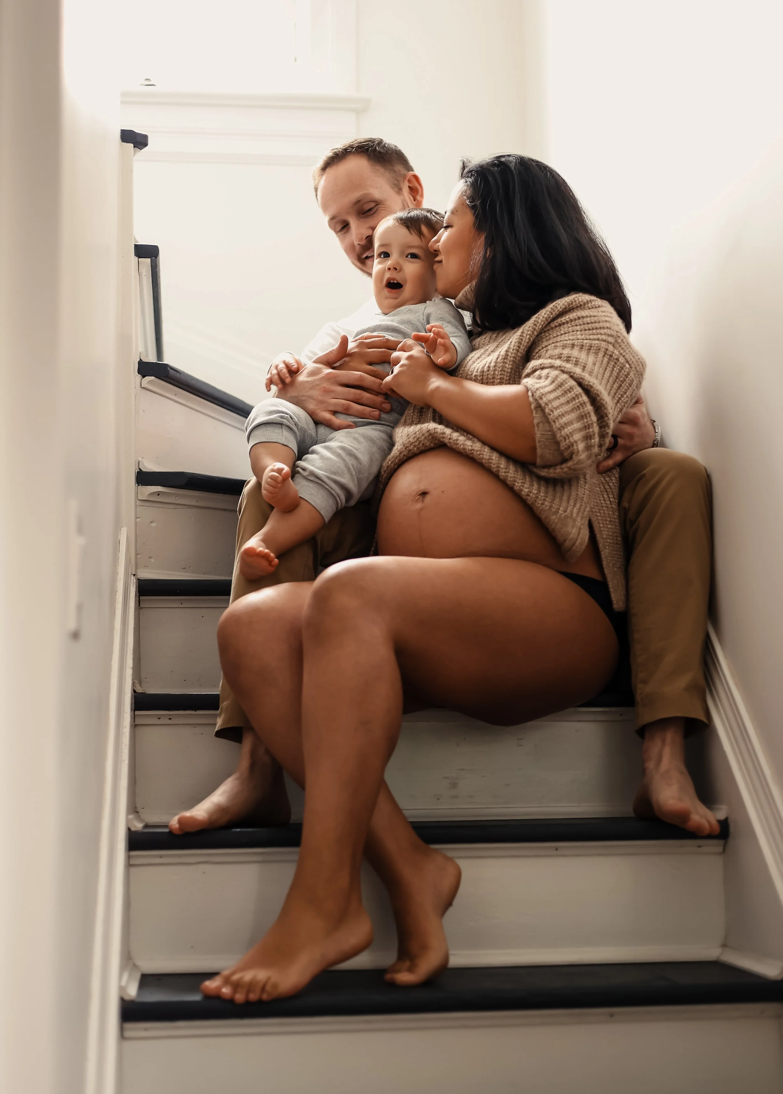 A pregnant woman sitting on stairs with her legs crossed, embracing a toddler boy while a man, possibly her partner, sits behind her. The woman is holding the toddler close, and they are all smiling and interacting affectionately.