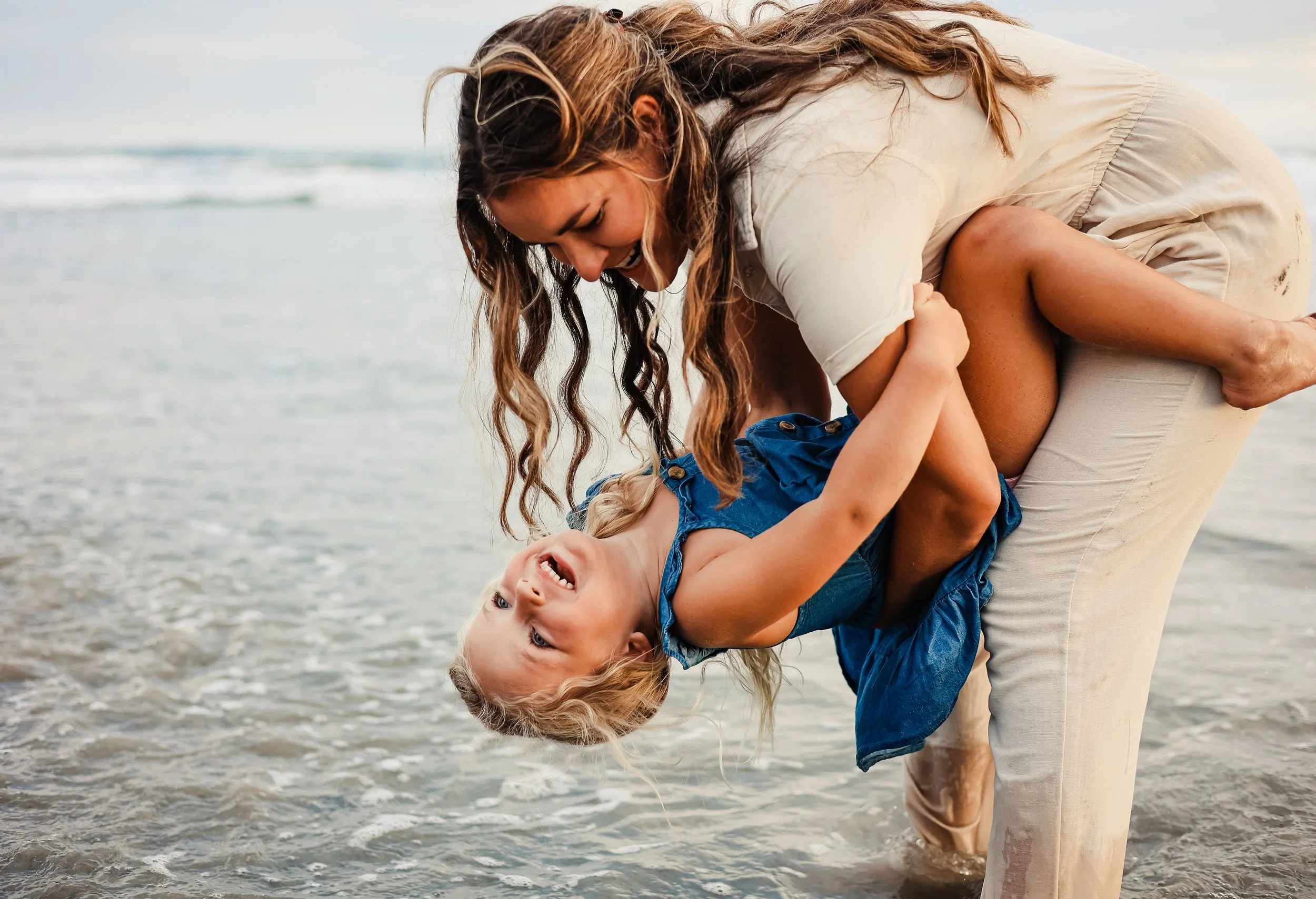 A woman is lifting a young girl into the air and they are playing at the beach by the water, smiling and enjoying themselves.