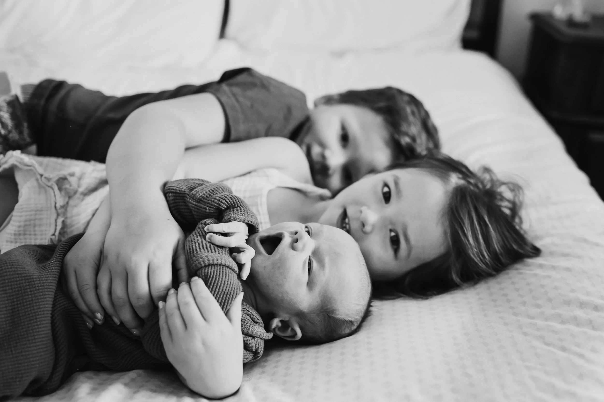 Three children lying on a bed, with two older ones gently holding a baby, all smiling and close together.