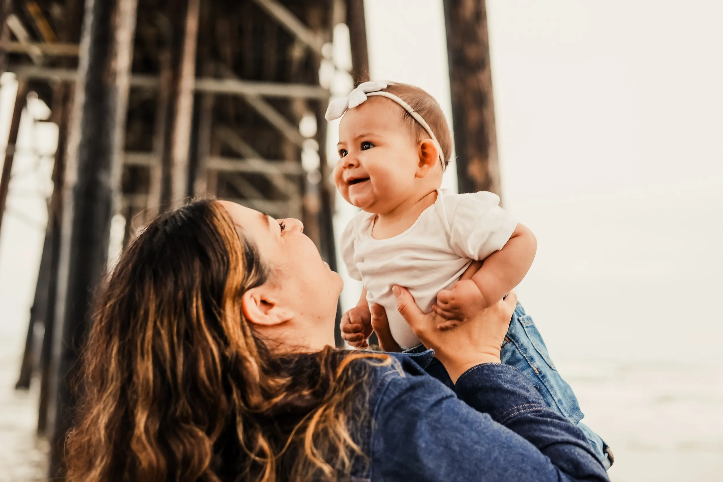 A woman with long, wavy hair holding a smiling baby girl up in the air under a wooden pier at the beach.