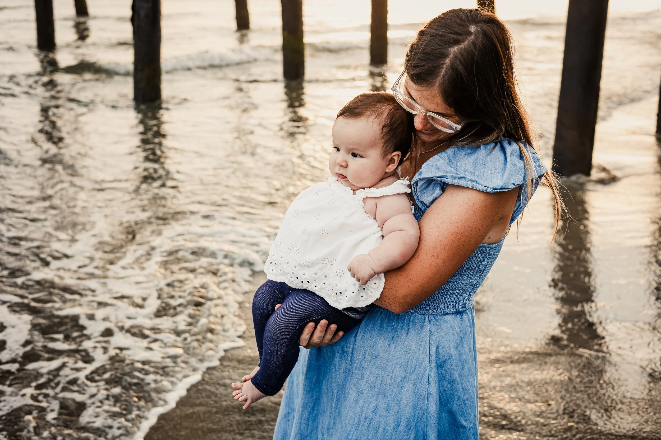A woman holding a baby girl by the beach at sunset with wooden pier posts in the background.