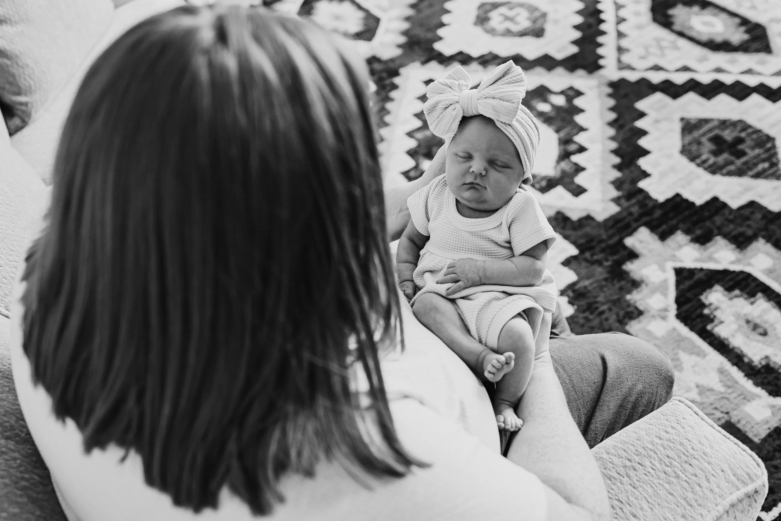 A woman holding a sleeping baby girl with a bow on her head, sitting on a patterned rug.