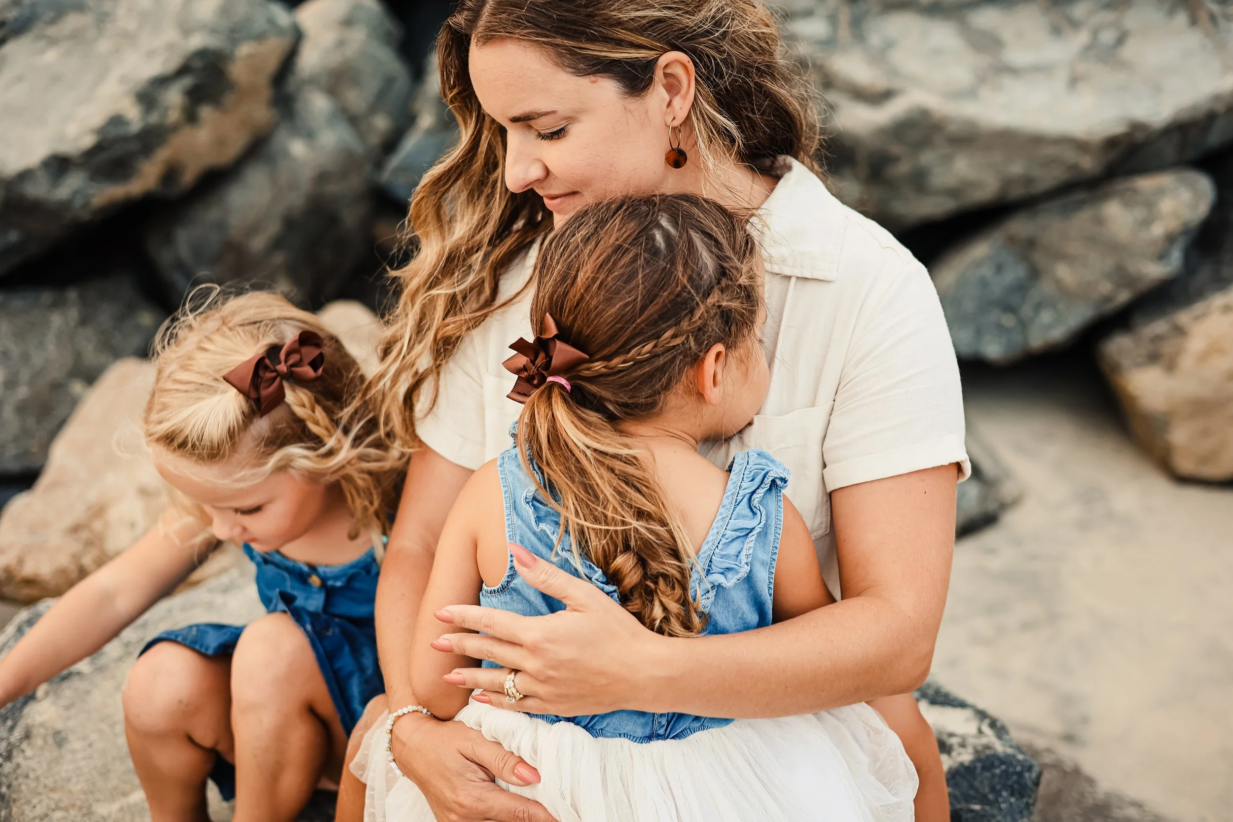 A woman with three young girls sitting on rocks at the beach, embracing one girl while the other girl looks down.