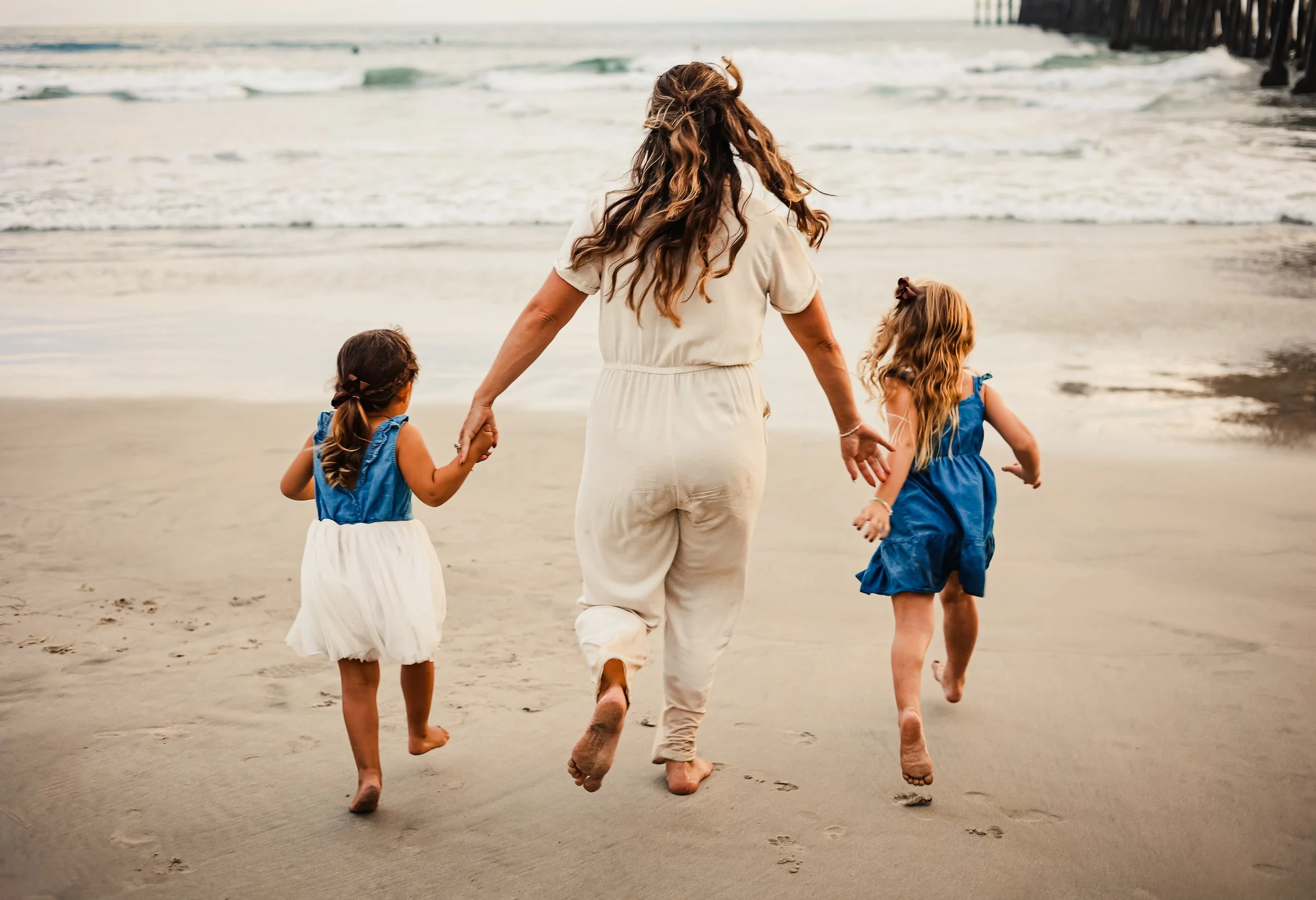 A woman and two young girls walking barefoot on the beach, holding hands, near the shoreline with waves and a pier or structure in the background