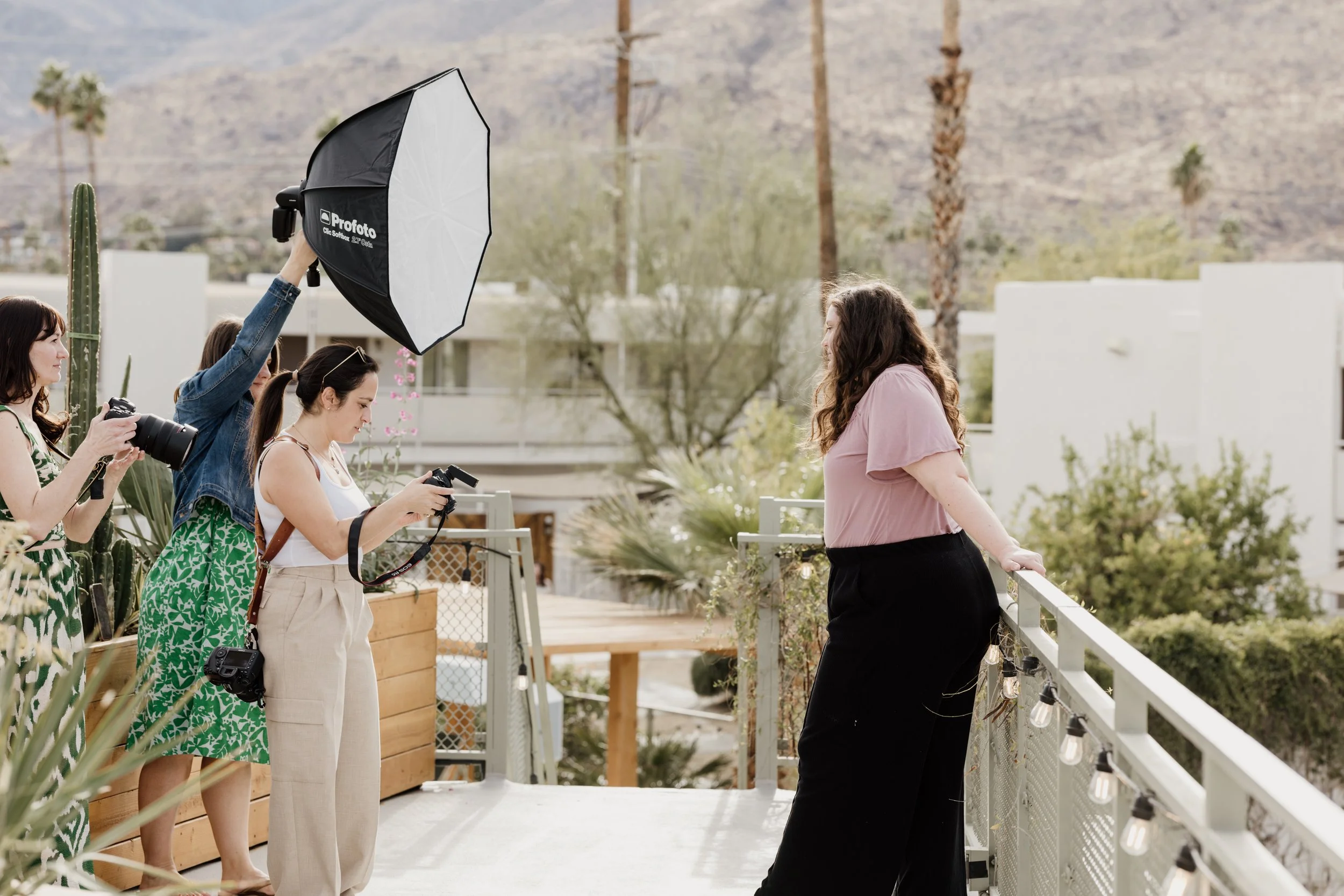 A woman standing on a balcony posing for a photo being taken by three women with cameras and a large lighting umbrella. The background shows a desert landscape with mountains, palm trees, and modern buildings.