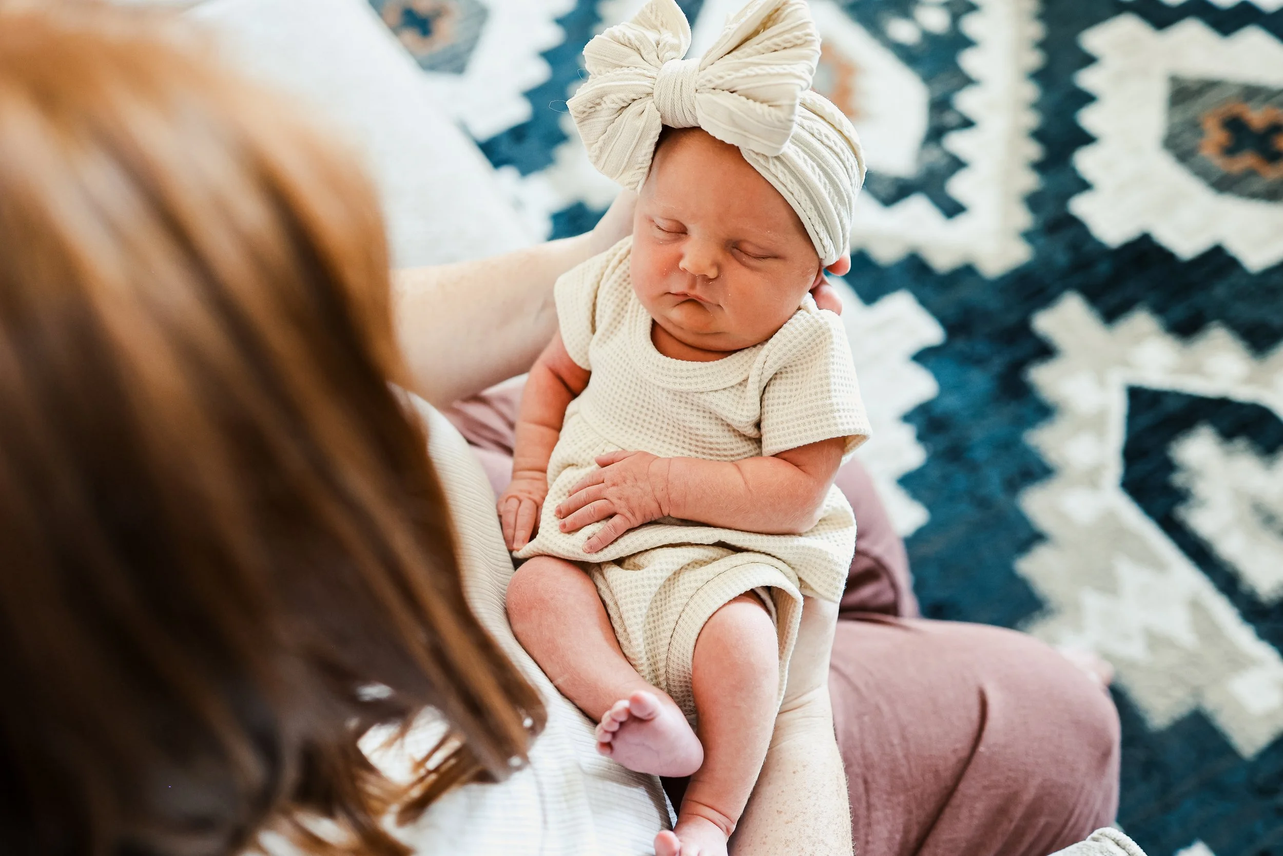 A baby wearing a cream-colored outfit and a headwrap is being held on a person's lap, with a patterned rug in the background.