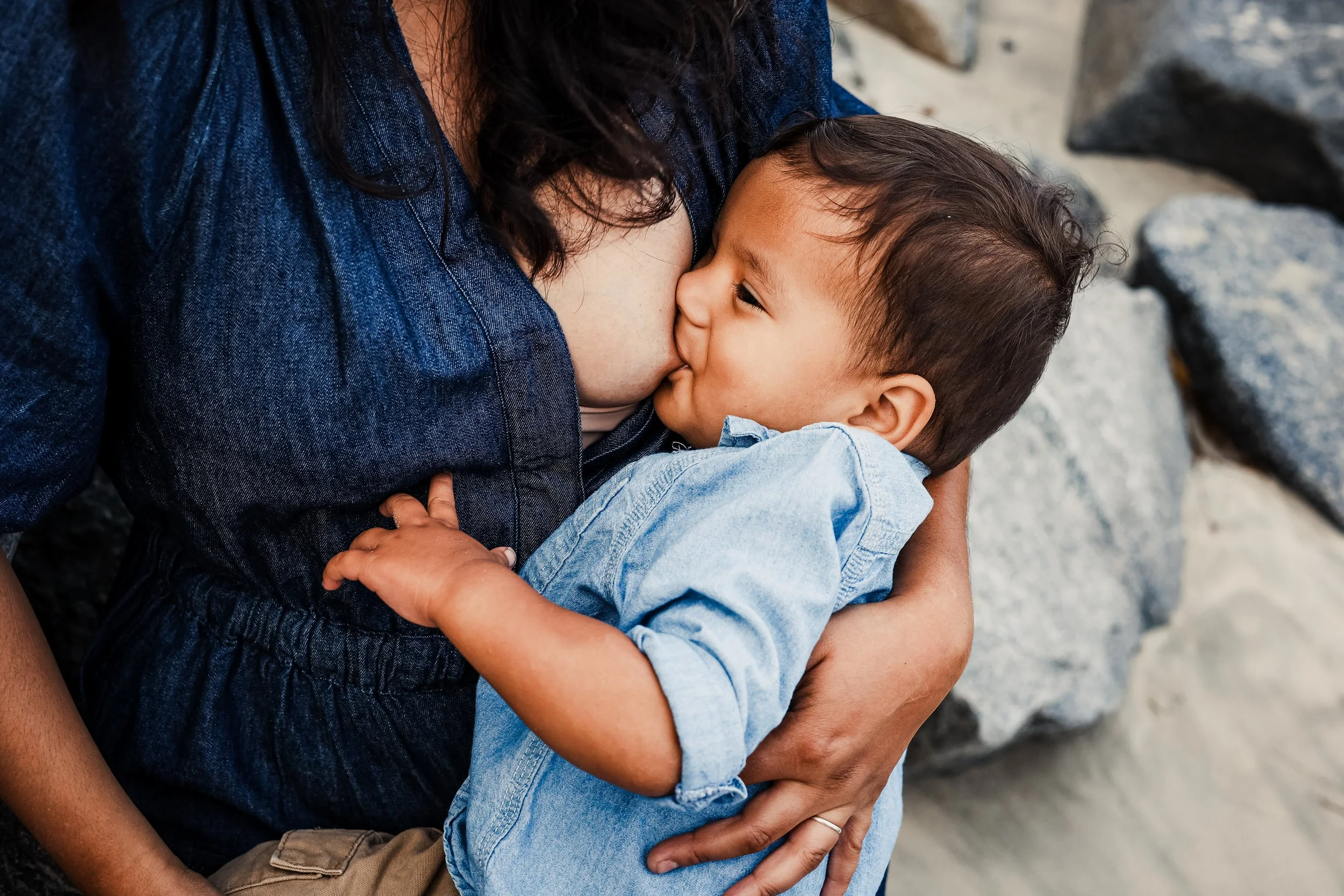 A young boy being breastfed by a woman, likely his mother, on a rocky beach.