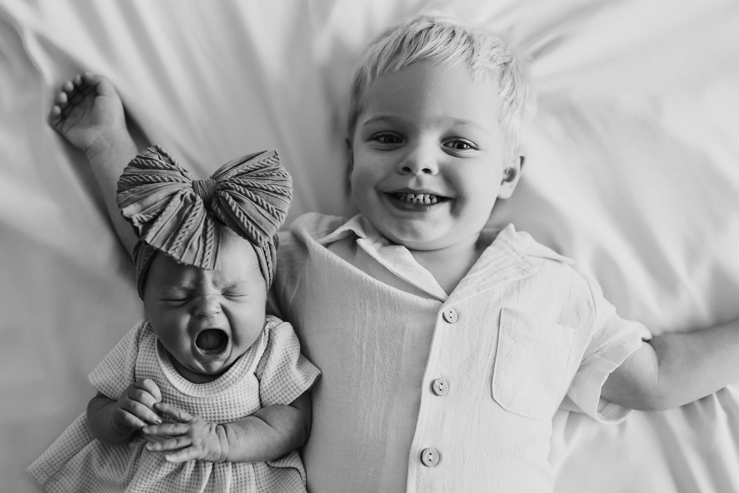 A young boy lying on a bed smiling and showing his teeth next to a crying baby girl with a large bow on her head.