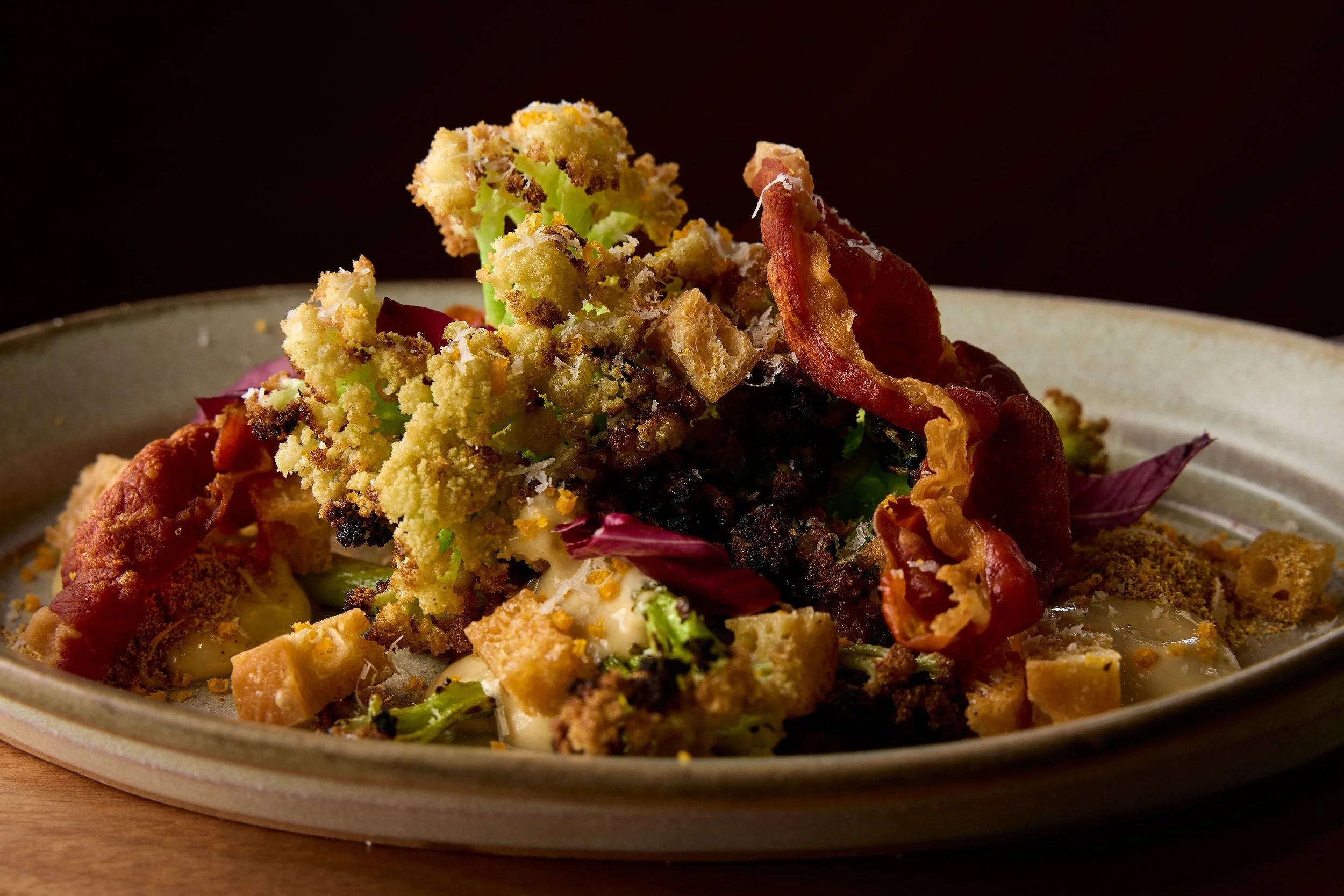 Close-up of a plate piled with diverse food items, including crispy bacon, bread crumbs, and greens, with a dark background.