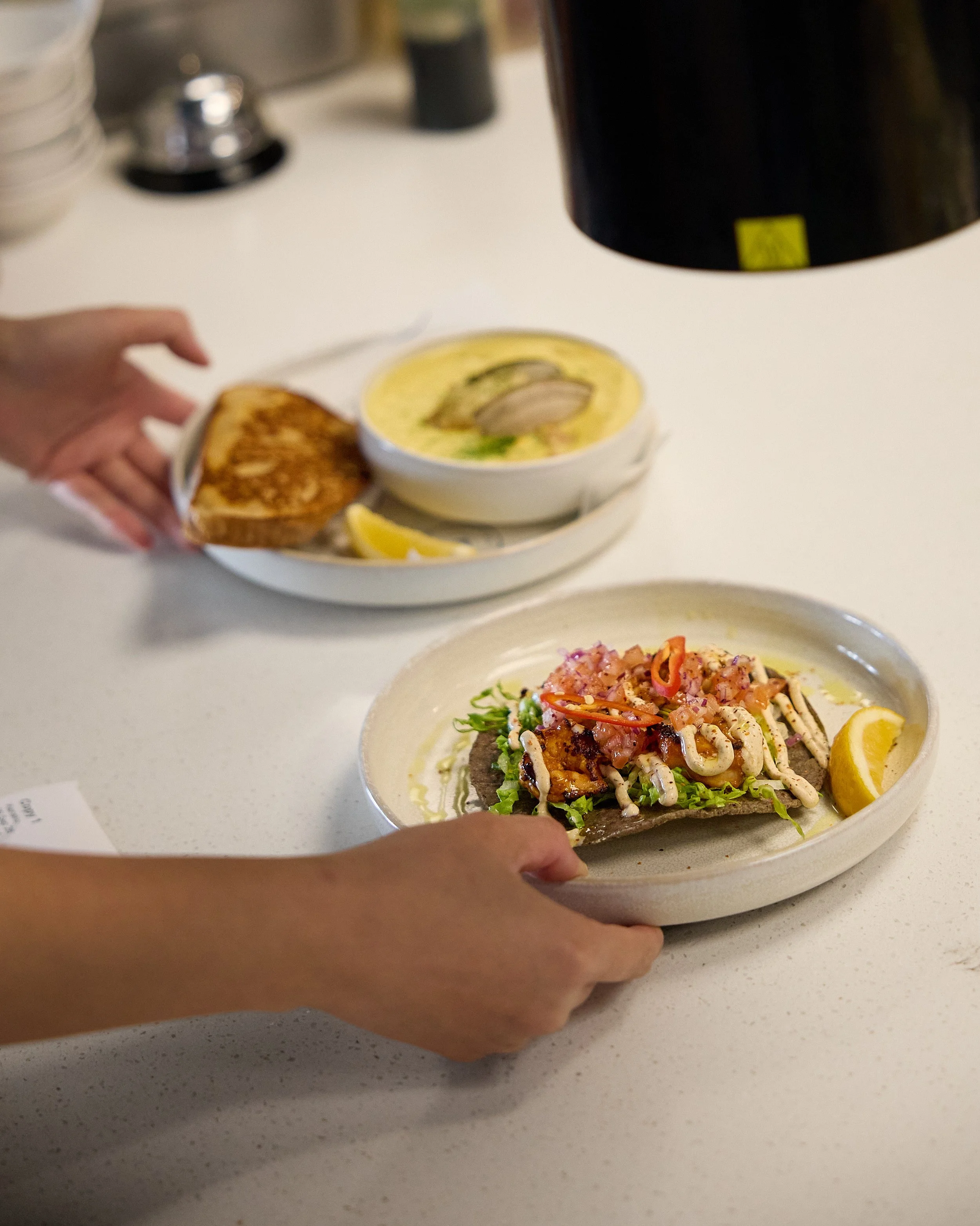 A hand holding a plate with loaded nachos topped with shredded cheese, diced tomatoes, sour cream, and a lemon wedge. In the background, a bowl of soup with garnish and a toasted bread slice are visible on a white counter.