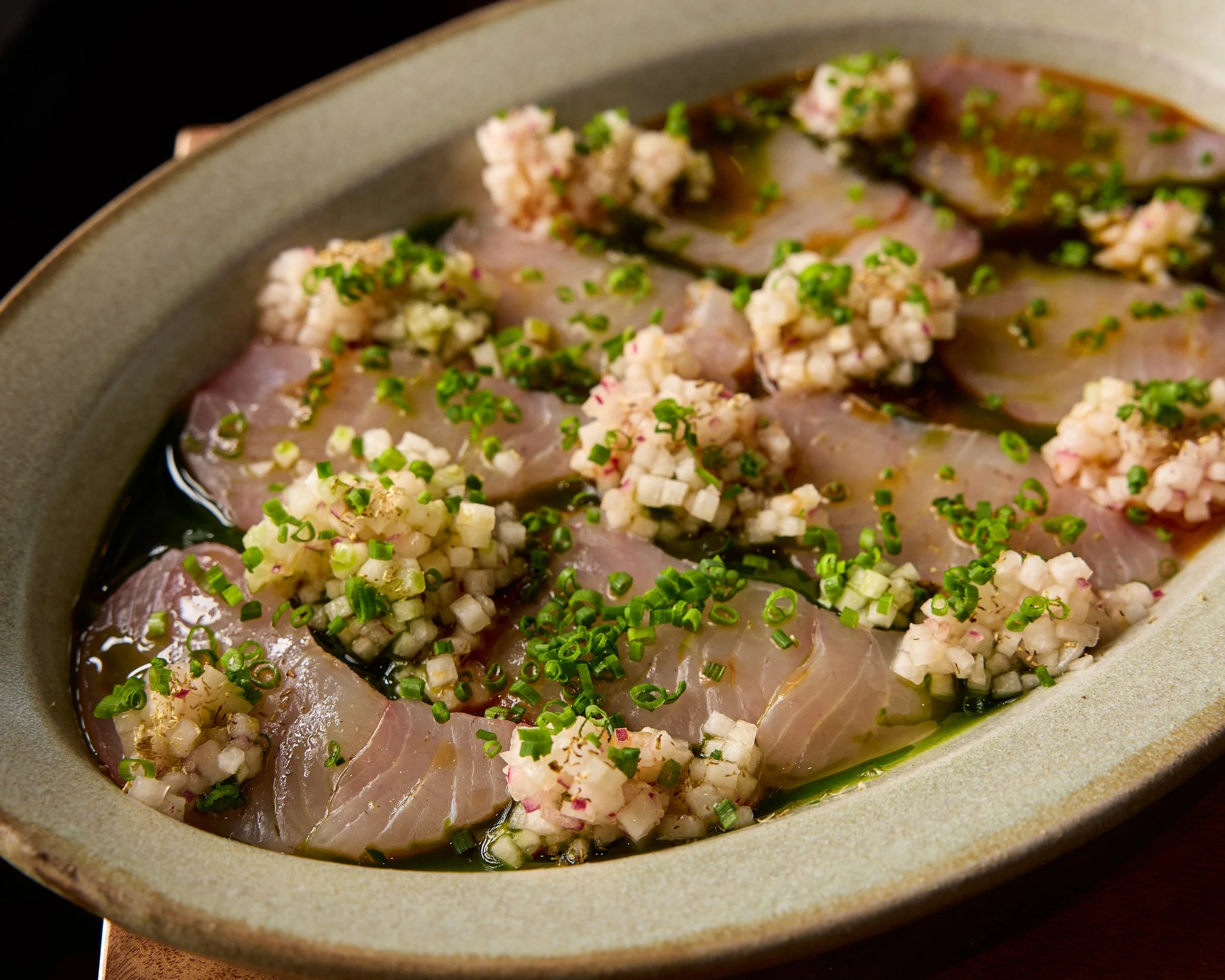 Sliced fish topped with chopped green onions and finely diced radishes in a shallow bowl with broth.