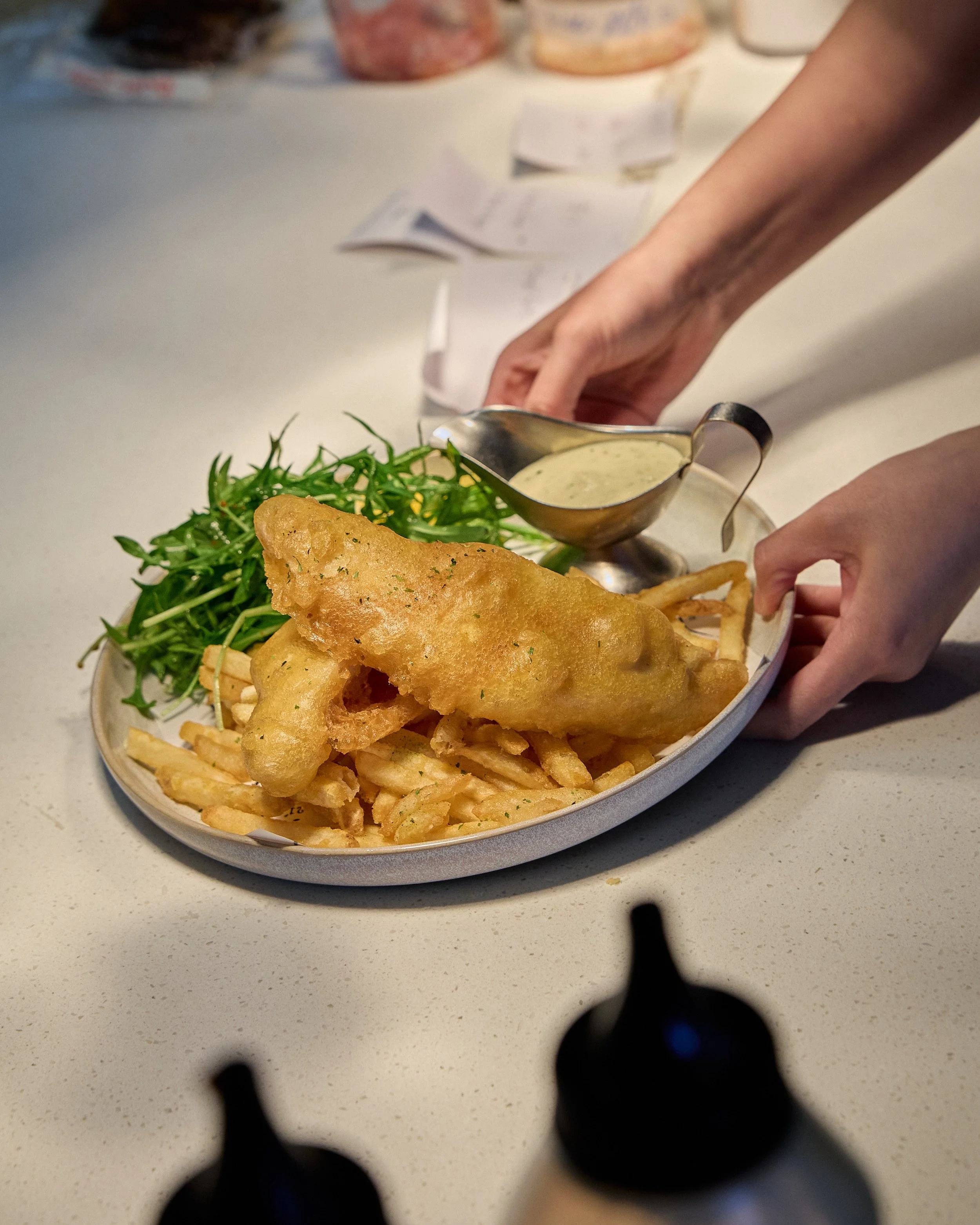 A plate of fish and chips with a side of greens and a small container of tartar sauce, seen from underneath a chess piece.