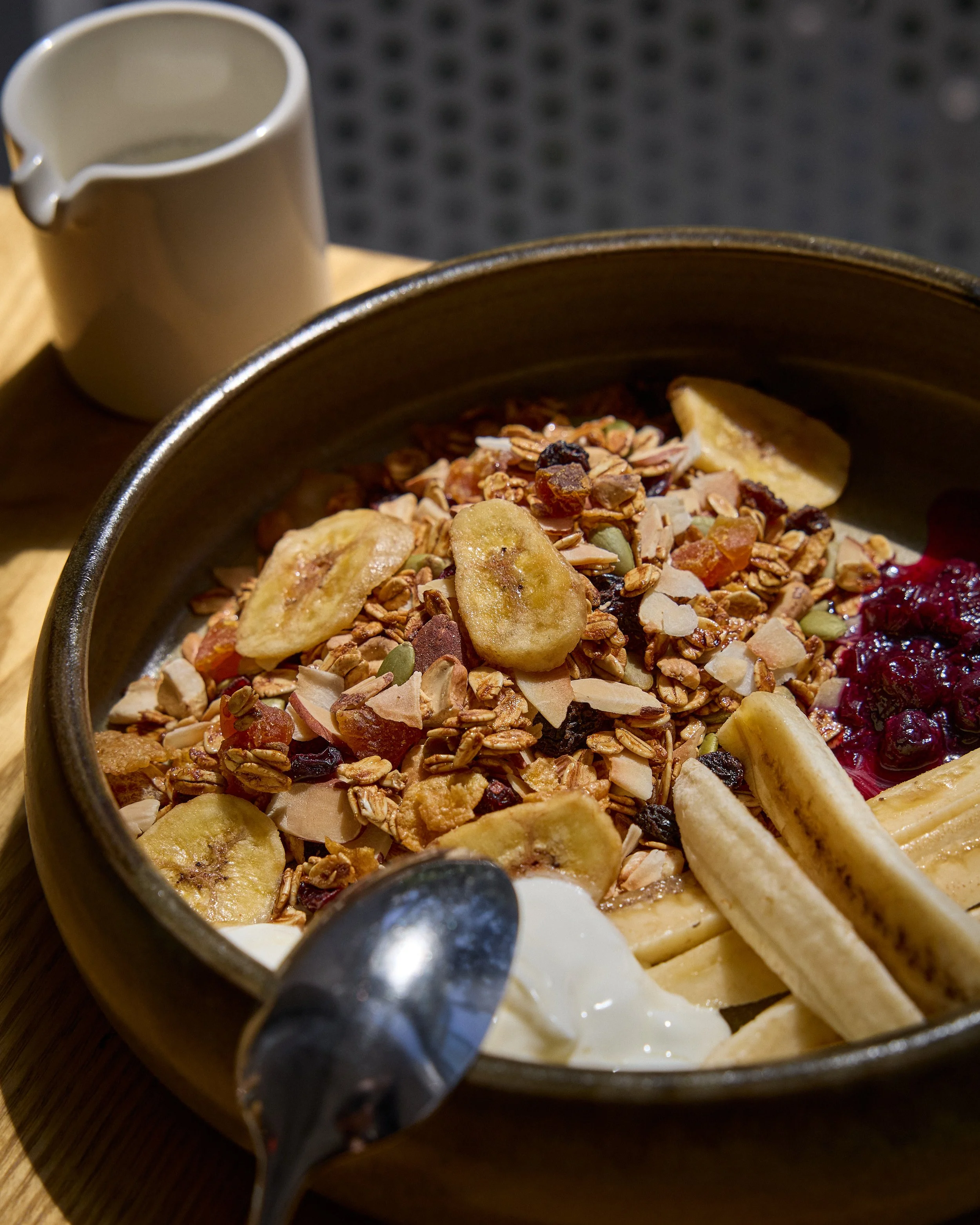 Close-up view of a bowl of yogurt topped with granola and banana slices, with a small pitcher of cream or milk in the background.