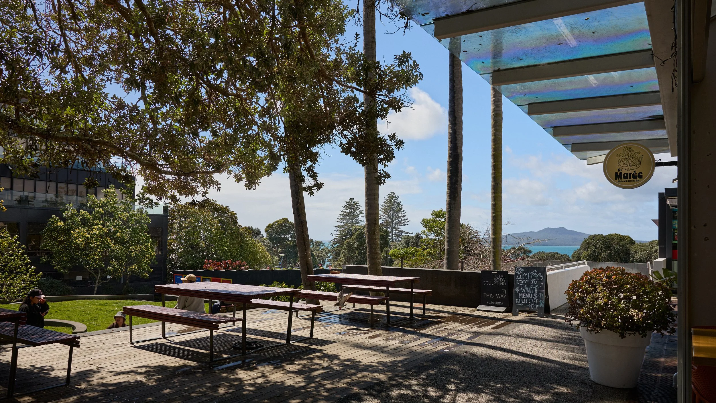 Outdoor patio with wooden tables and benches under trees, sign for 'S. Marée Bistro & Coffee Bar', view of ocean and distant islands in the background.