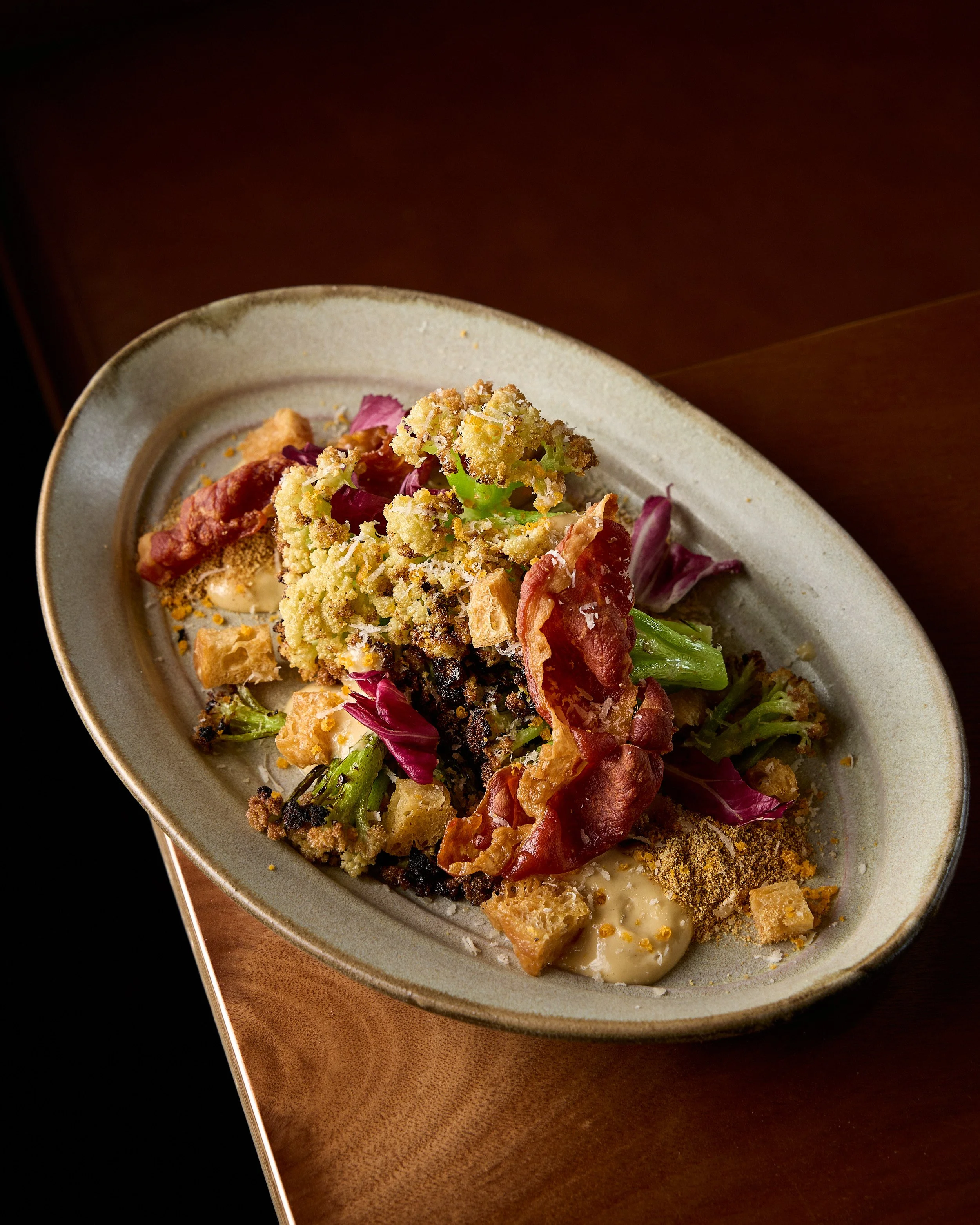 Close-up of a gourmet appetizer on a ceramic plate, including crispy bread crumbs, smoked bacon, leafy greens, and crumbled cheese, on a wooden table.