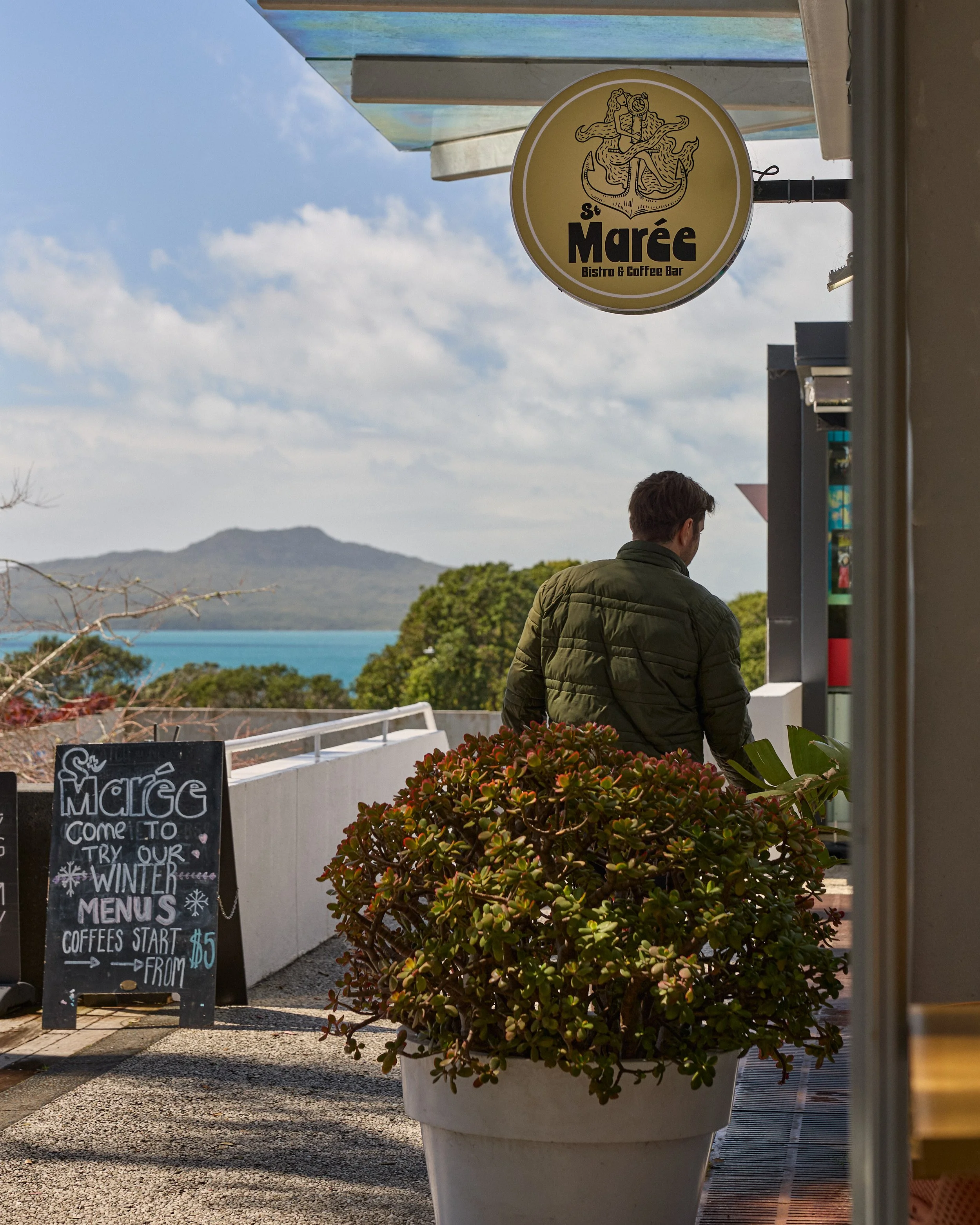 Sign for St. Marée Bistro & Coffee Bar with a logo of a mermaid holding a trident, against a blue and cloudy sky. Part of a person in a green jacket is visible, along with a large potted plant in the foreground and a blackboard sign advertising winte