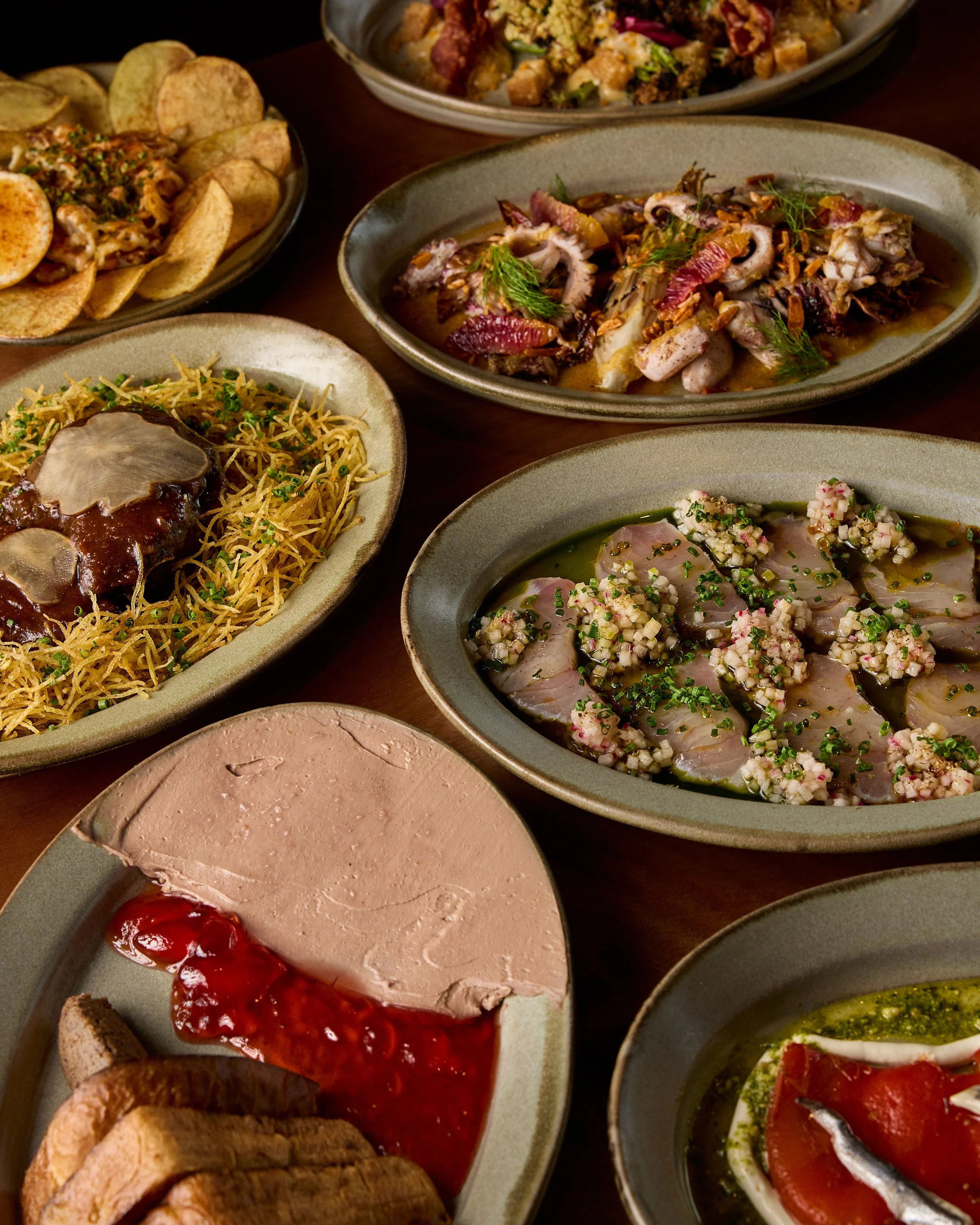 Assorted dishes of appetizers and snacks on a table, including sliced meats with herbs, potato chips, pasta with sauce, and vegetables.