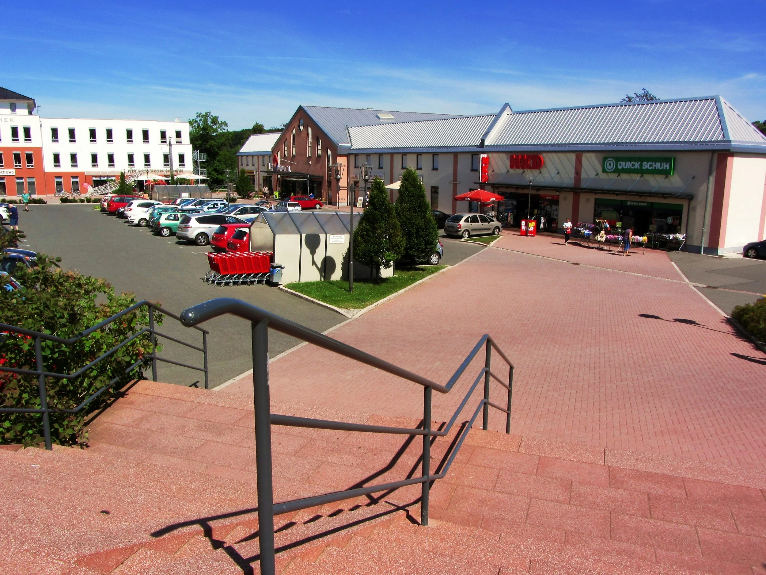 Shopping plaza with parking lot, storefronts, and a few people walking in sunny weather.