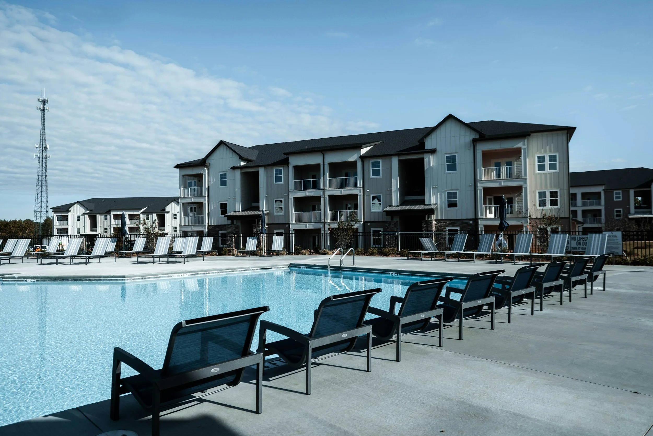 Empty outdoor swimming pool with lounge chairs and apartment buildings in the background under a partly cloudy sky.