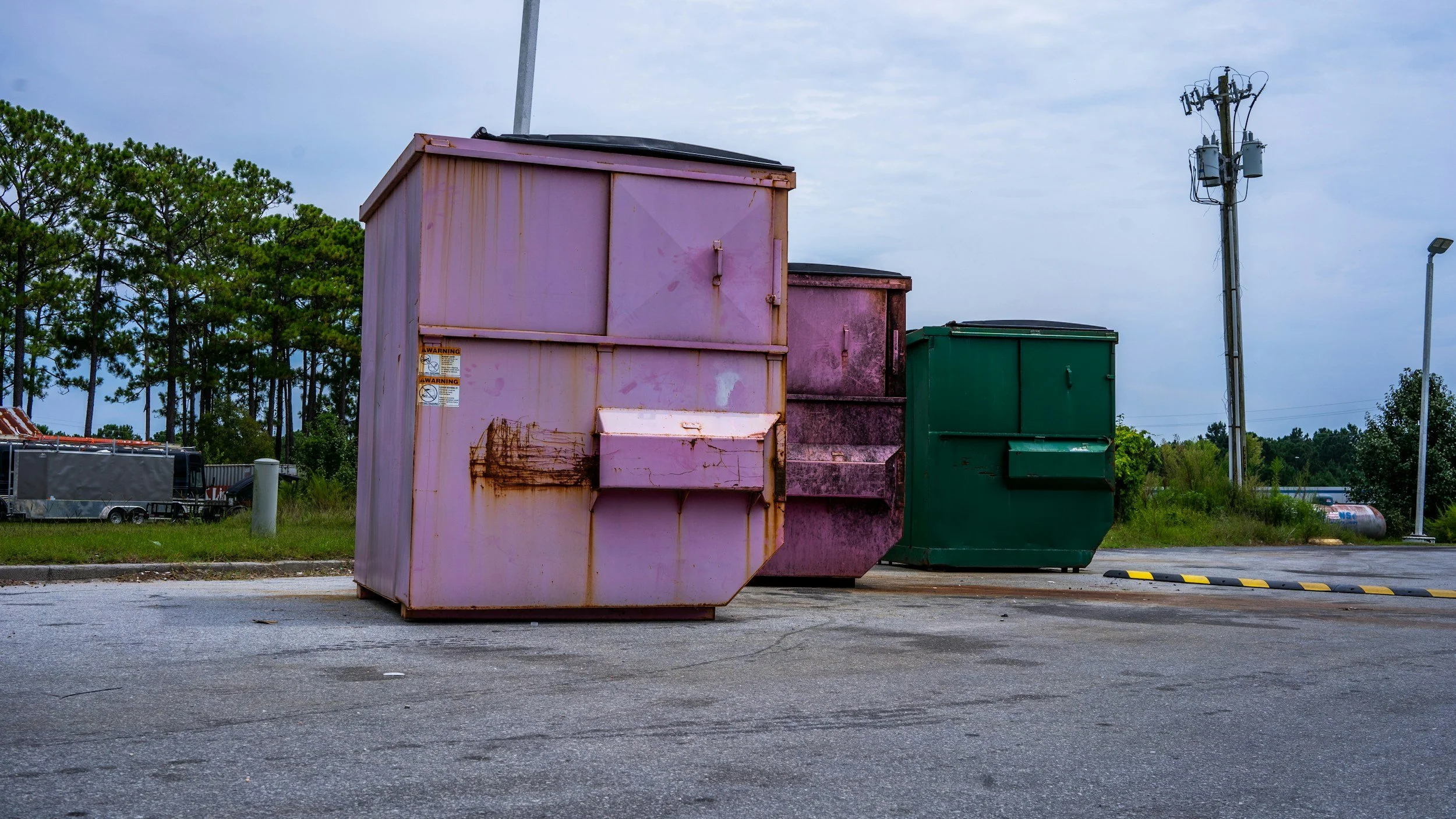 Three large garbage dumpsters in pink, purple, and green, placed on a paved lot with a grassy background and a row of trees, under a cloudy sky.