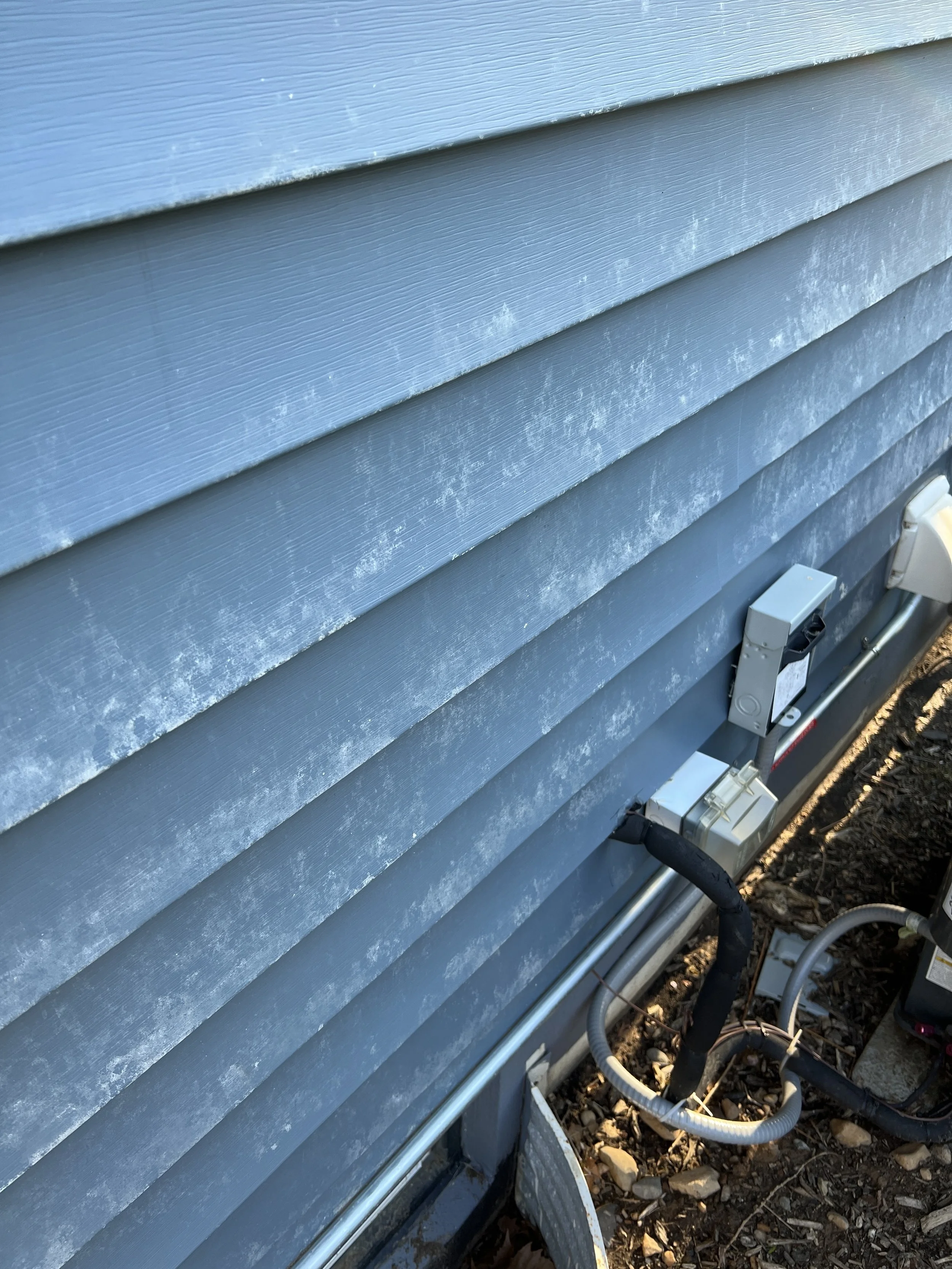 A close-up of the side of a house with grey horizontal siding, showing white mold or mildew on the exterior wall, along with electrical boxes and conduit pipes at the bottom, and a patch of soil with small rocks.