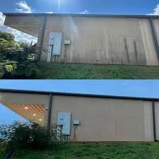 Side view of a beige building with a flat roof, electrical boxes on the wall, and a grassy area with bushes in front.
