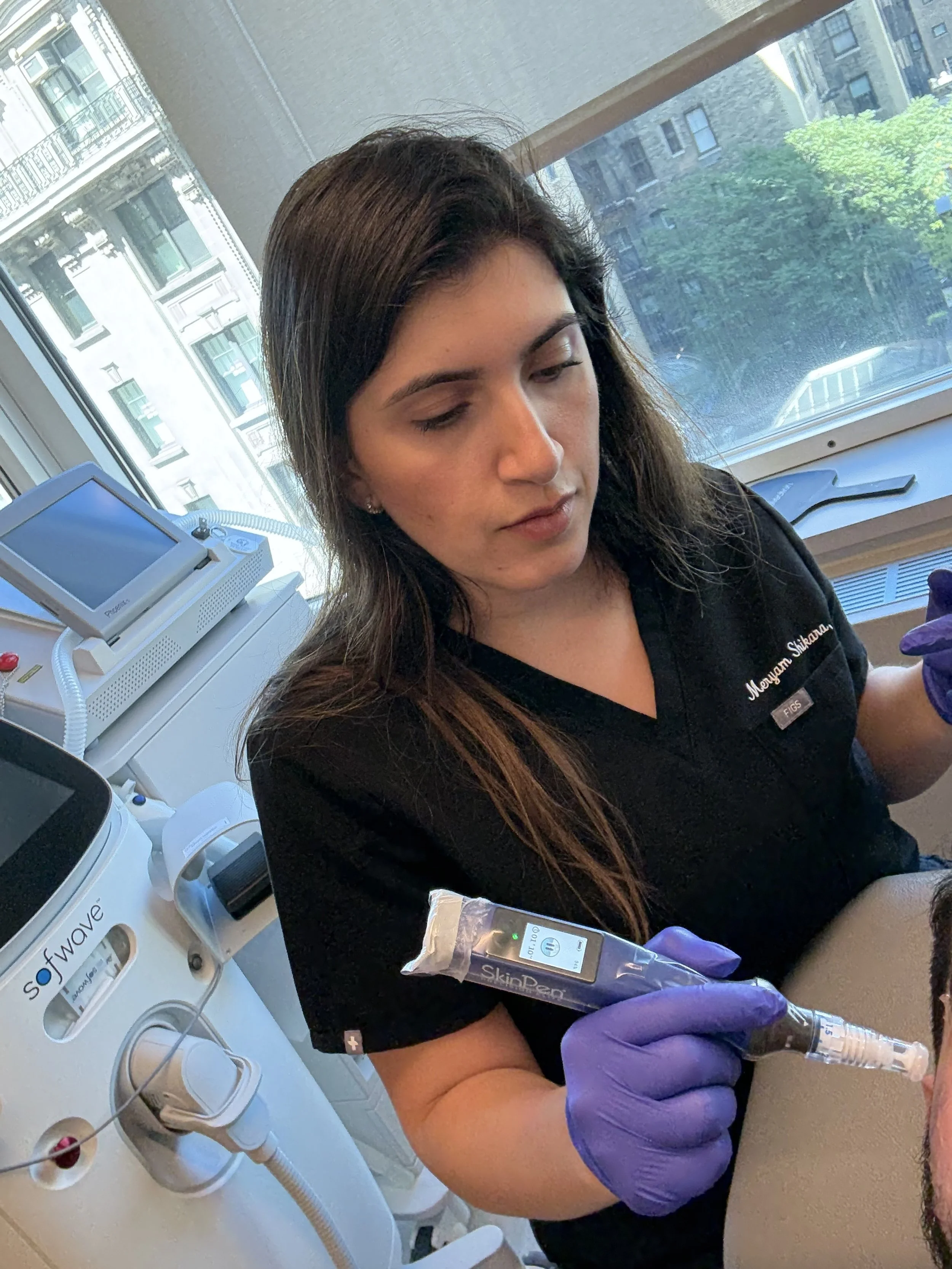 Meryam Shikara, MD a facial plastic surgeon in black scrubs is wearing purple gloves is administering microneedling to a patient's face in a medical clinic with large windows showing city buildings and trees outside.