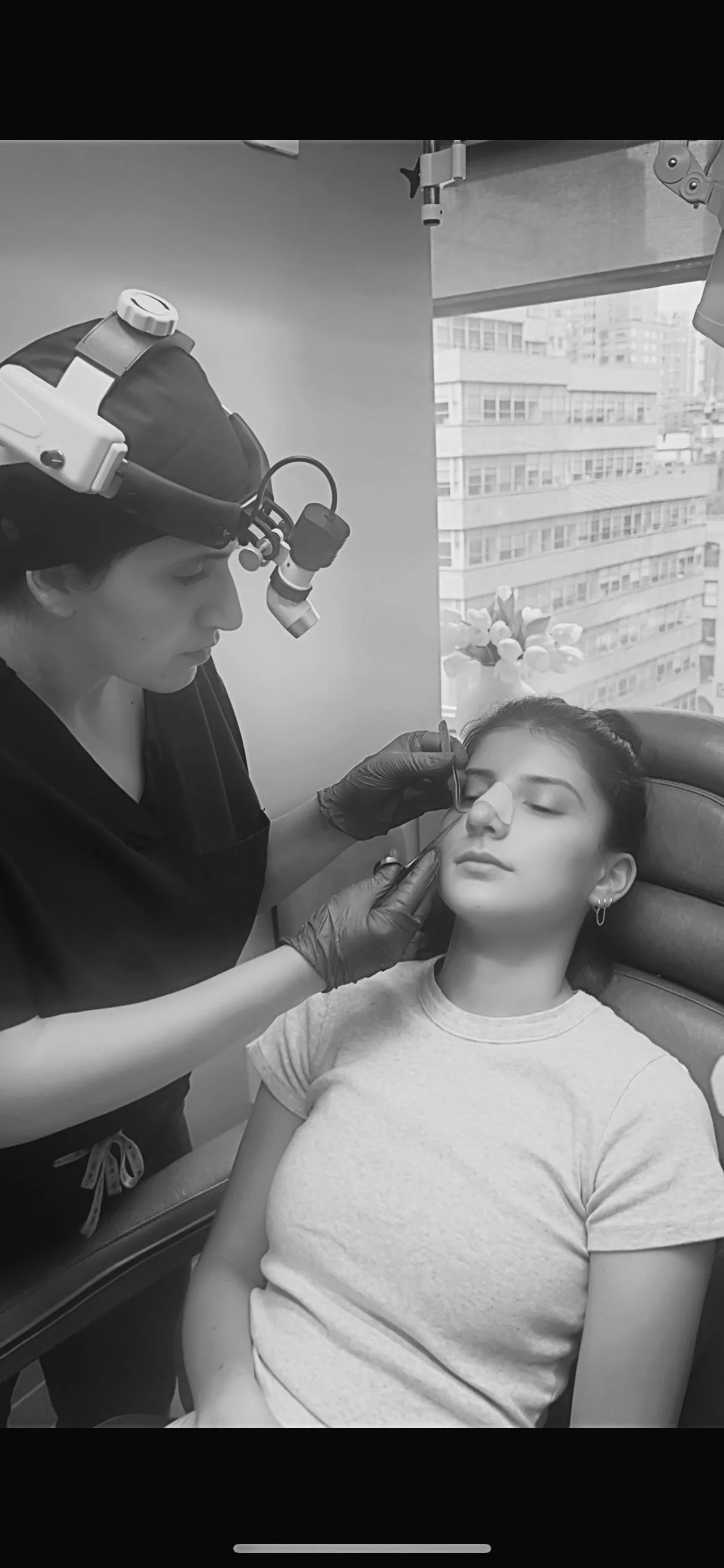A woman getting her cast removed after rhinoplasty surgery to improve breathing from the nose, cast is being removed by Meryam Shikara, MD, Facial plastic surgeon, in a clinical setting with a city view outside the window.