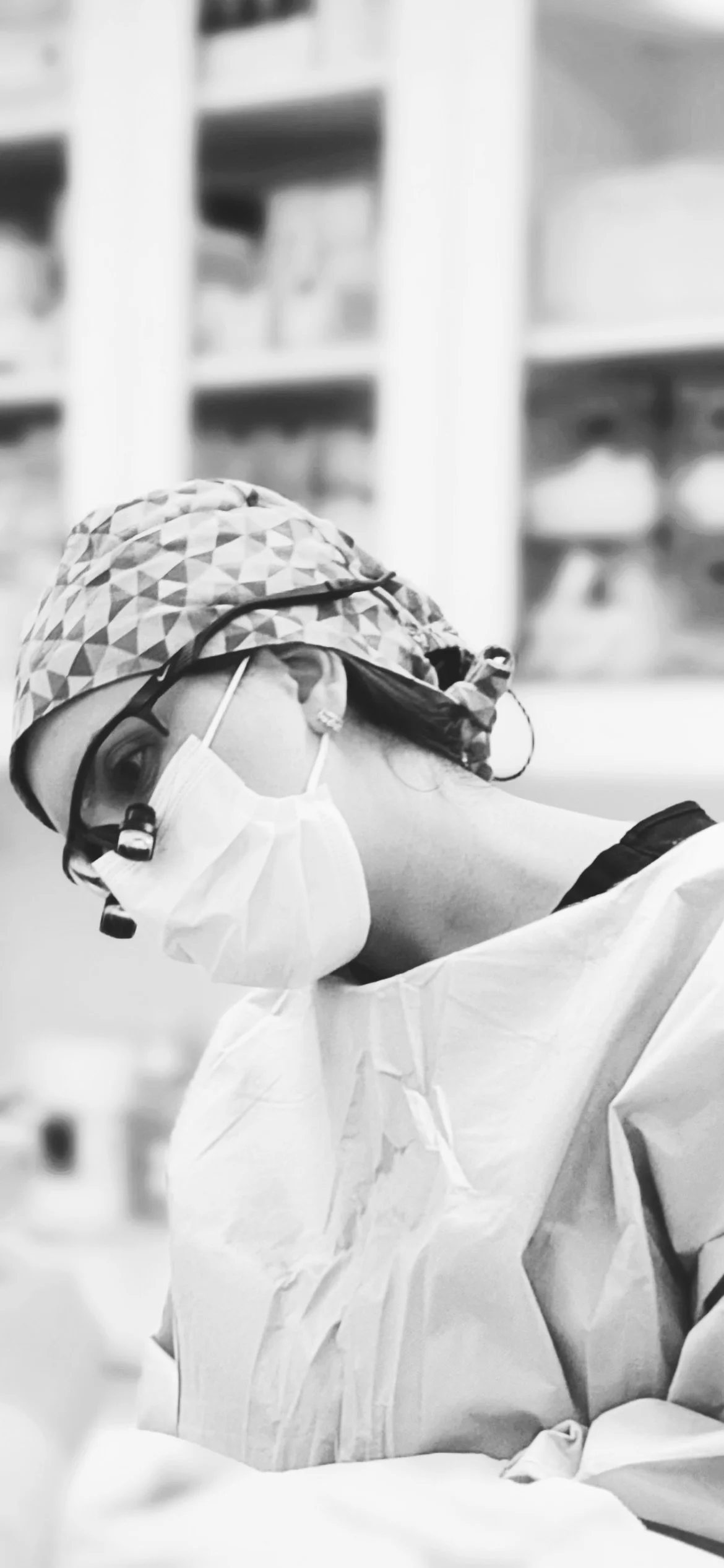 A black and white photo of a female healthcare worker wearing a patterned surgical cap, face mask, protective eyewear, and a disposable gown, working in a medical setting.