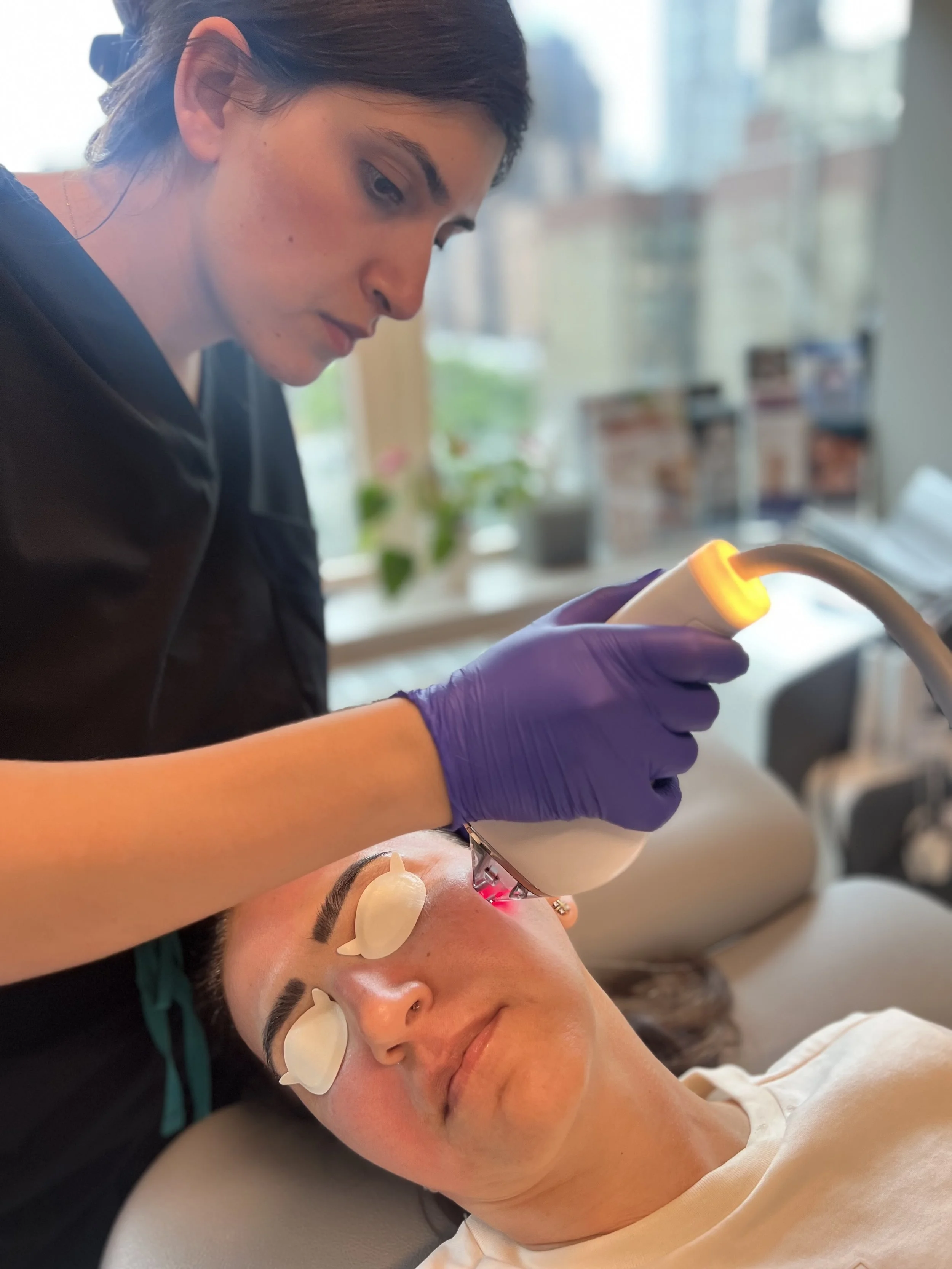 A woman receiving a laser facial treatment while lying on a medical bed in a clinic, with eye shields on her eyes and Meryam Shikara, MD a facial plastic surgeon is using a laser device.