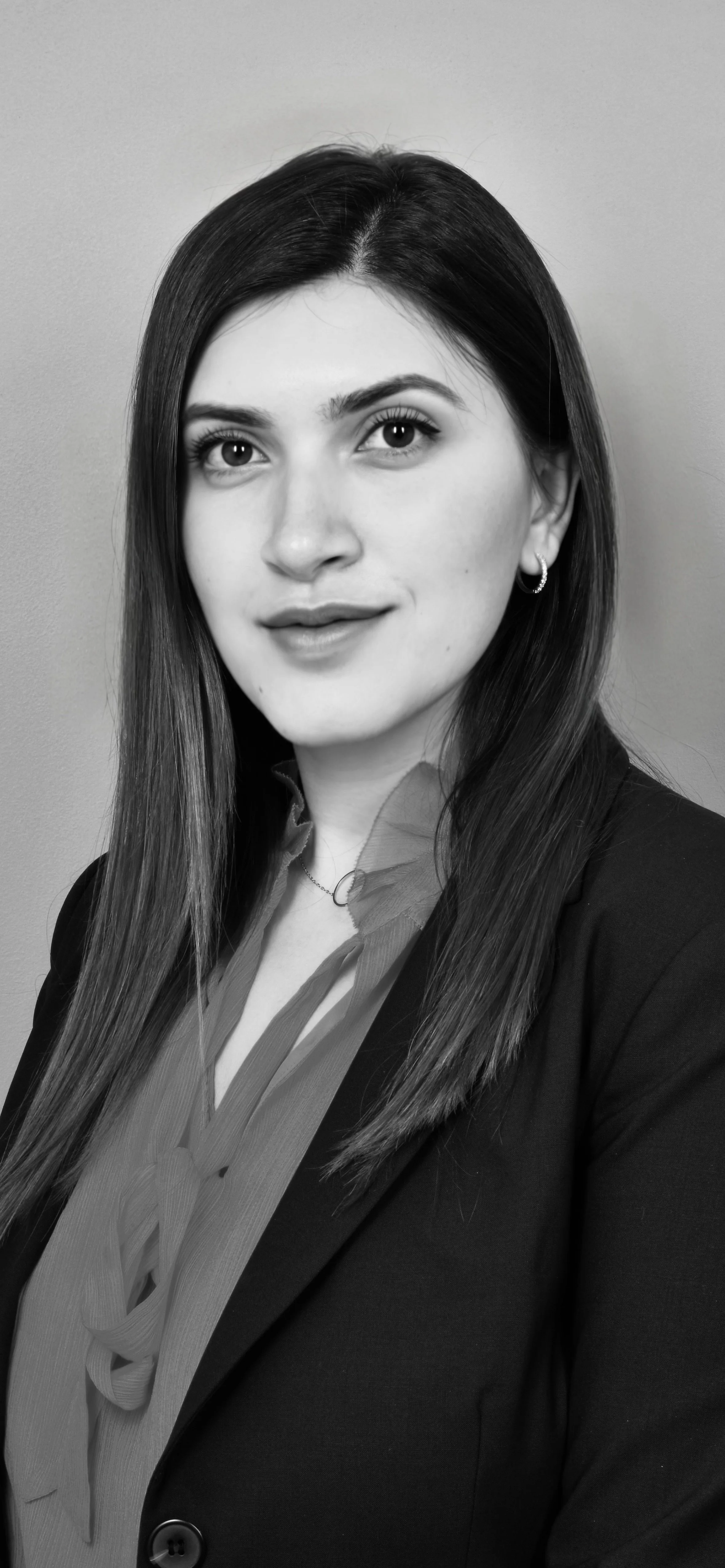 Black and white professional portrait of Dr. Meryam Shikara with long dark hair, wearing a blazer and blouse, standing against a plain background.