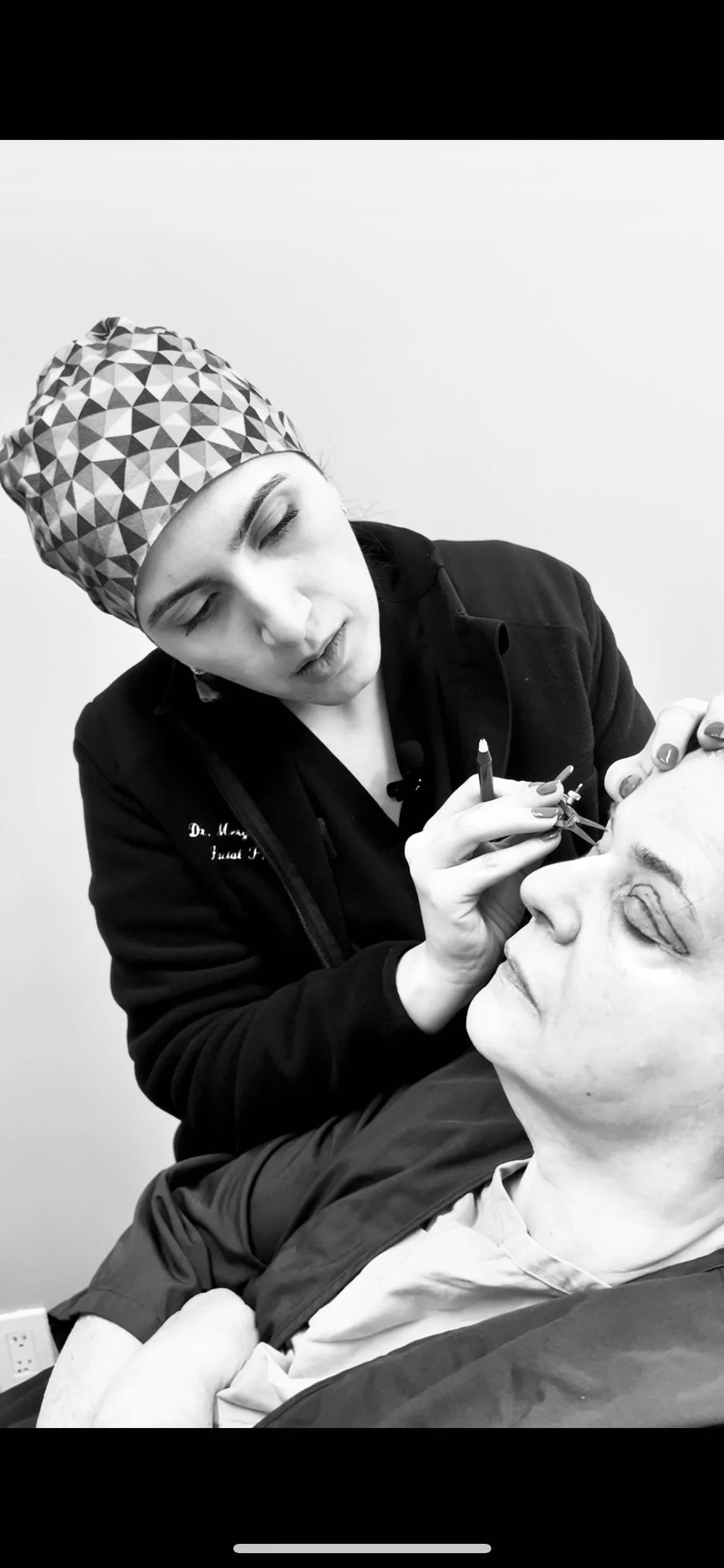 A woman is performing a cosmetic procedure on a woman lying down, using a pen-like instrument near her eye, in a clinical setting.