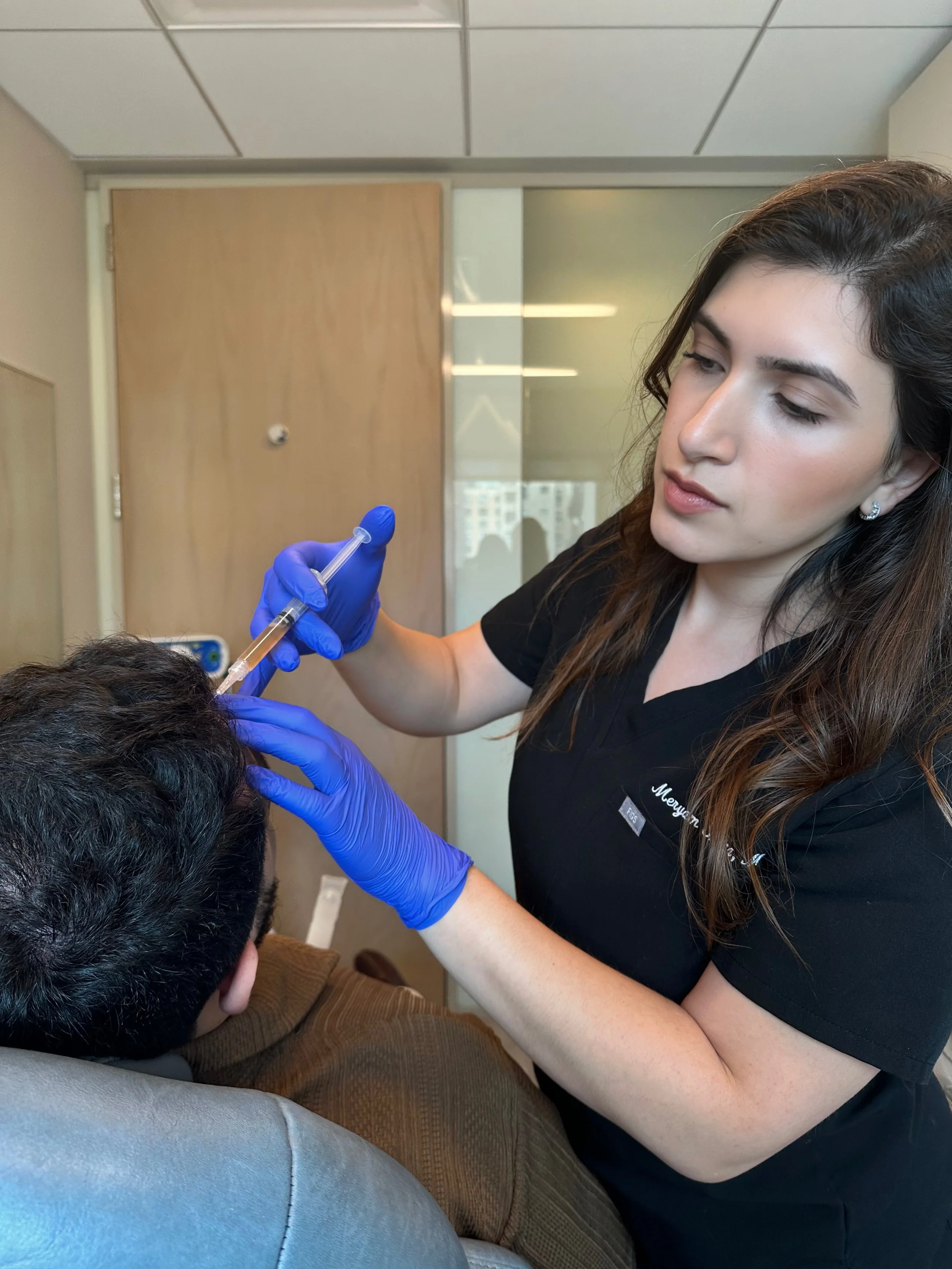 A female healthcare professional, Meryam Shikara, MD wearing blue gloves administering hair rejuvenation injection to a patient sitting in a chair.