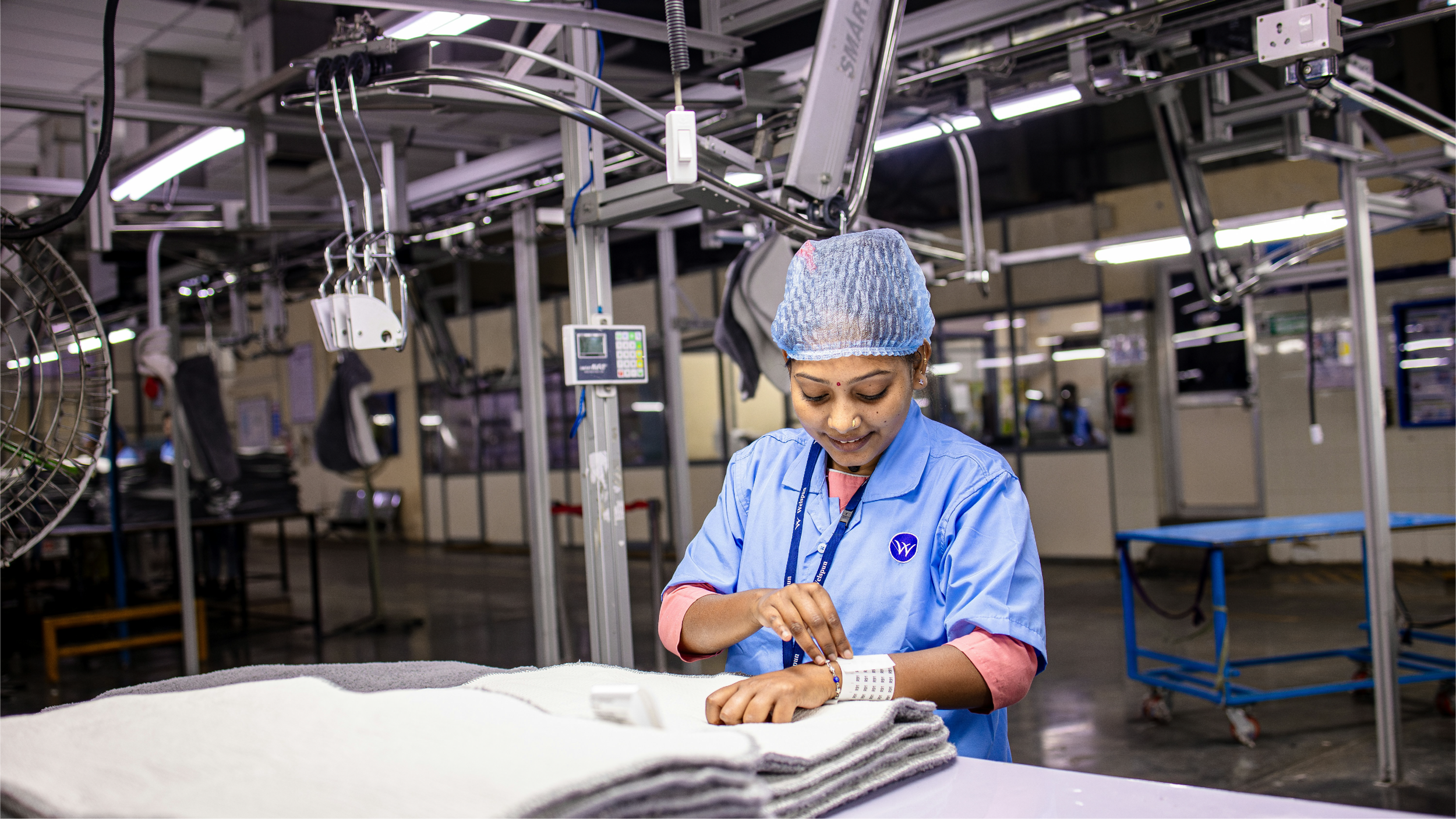 Female worker in a cleanroom, wearing a blue uniform, hairnet, and bracelet, inspects fabric or material in a manufacturing or industrial setting.