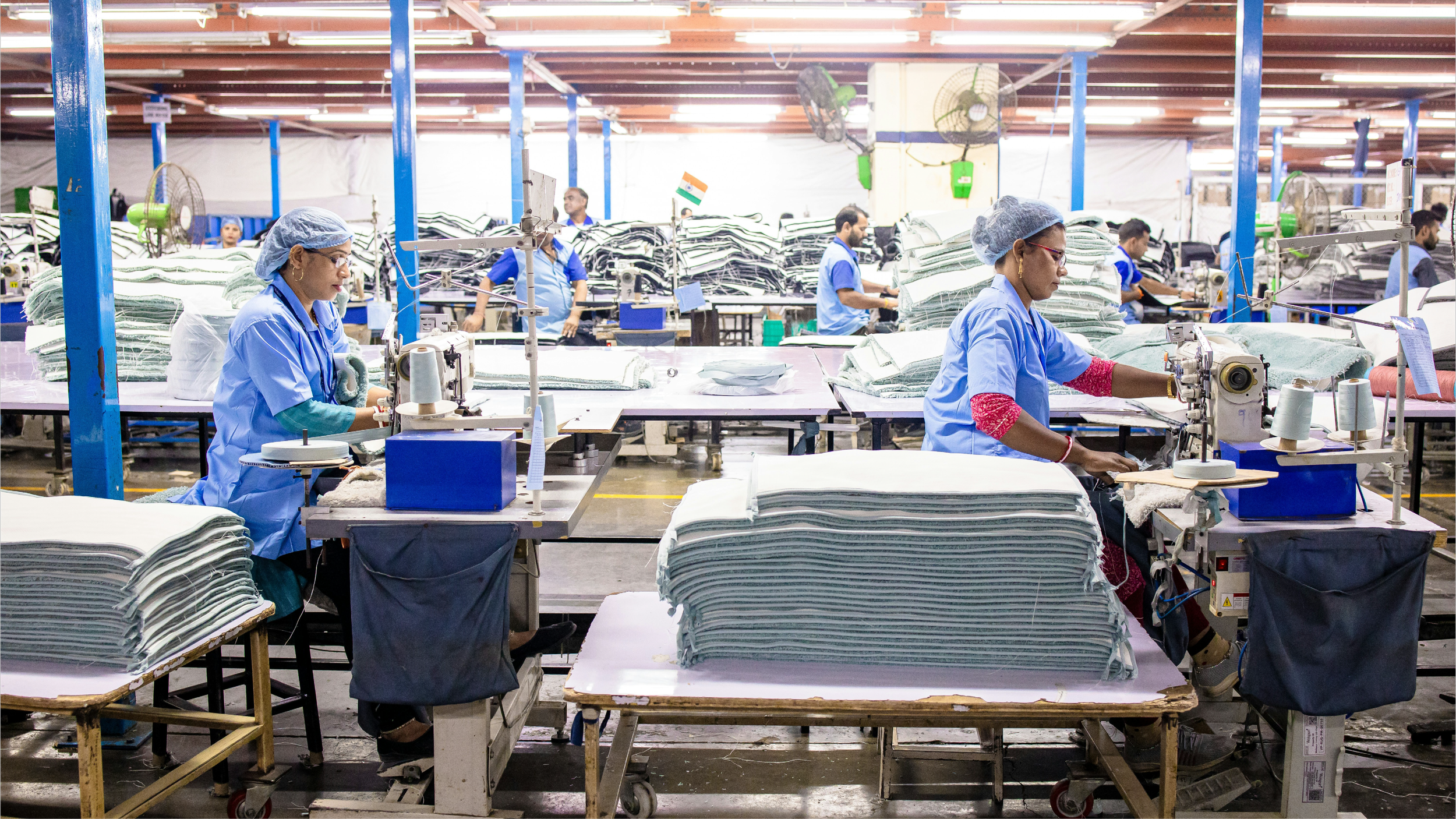 Workers in blue uniforms and hairnets sewing fabric in a textile factory with stacks of fabric and industrial machines.
