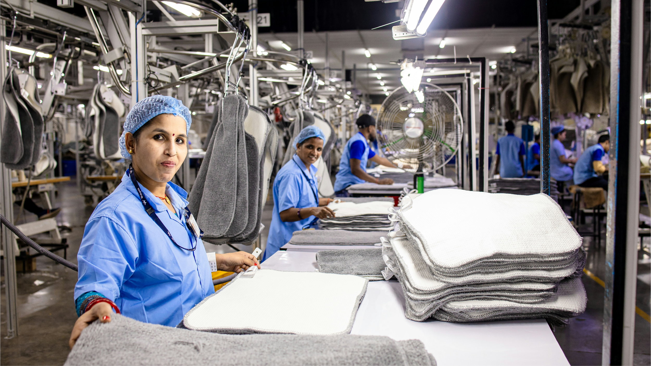 Workers in a factory assembling white and gray towels on a processing line with shelving and hanging towels in the background.