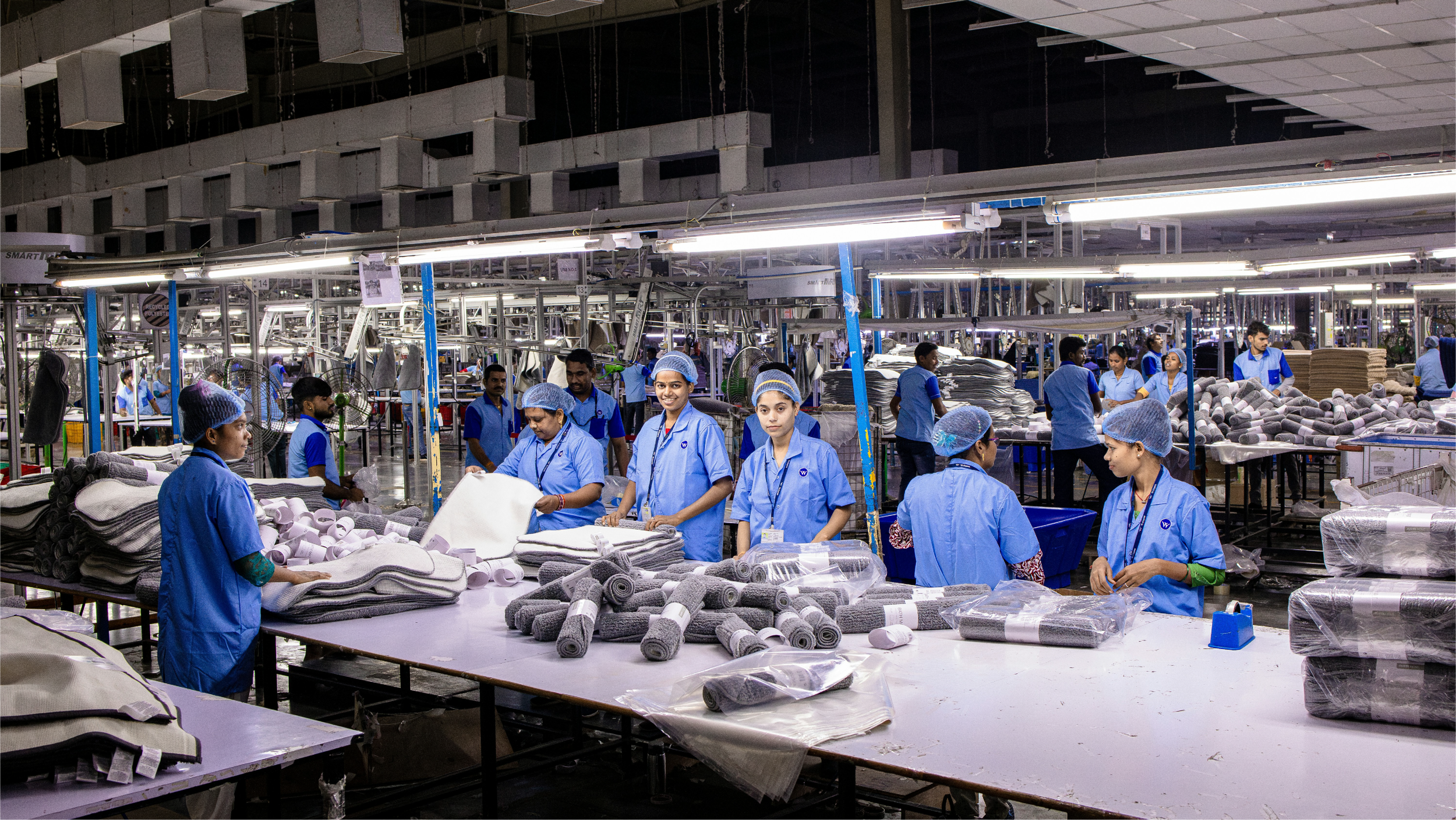 Workers in blue uniforms and hairnets work on textile or furniture product assembly in a large factory, with stacks of fabric and rolls of material on tables.