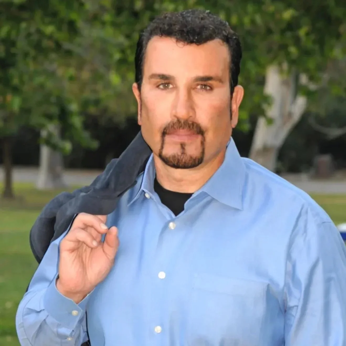 A man with dark hair, facial hair, wearing a light blue button-up shirt, standing outdoors with trees and grass in the background, holding a jacket over his shoulder.