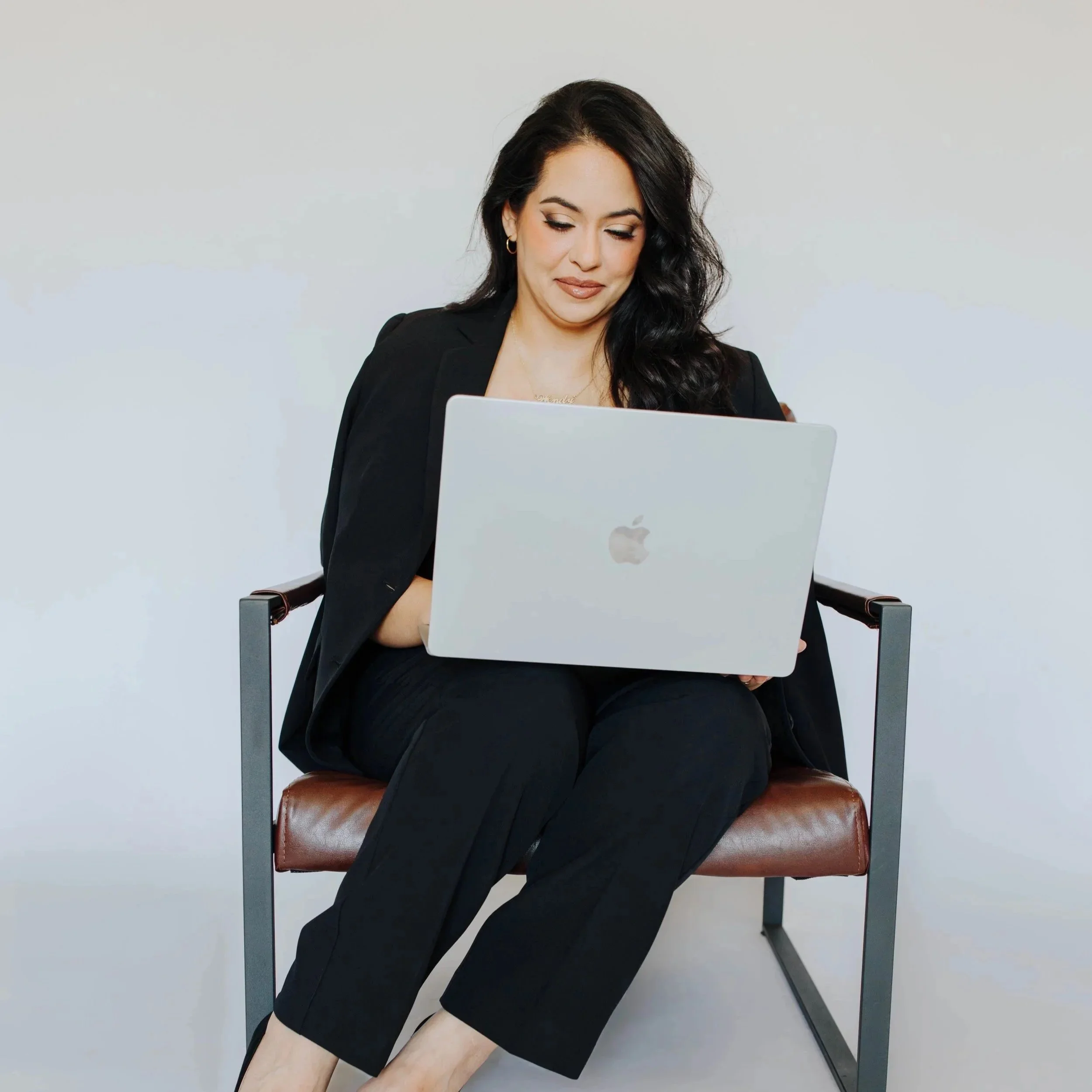 A woman sitting on a brown leather chair with a metal frame, using a silver MacBook laptop, dressed in black professional attire, with dark wavy hair, a subtle smile, and a white background.