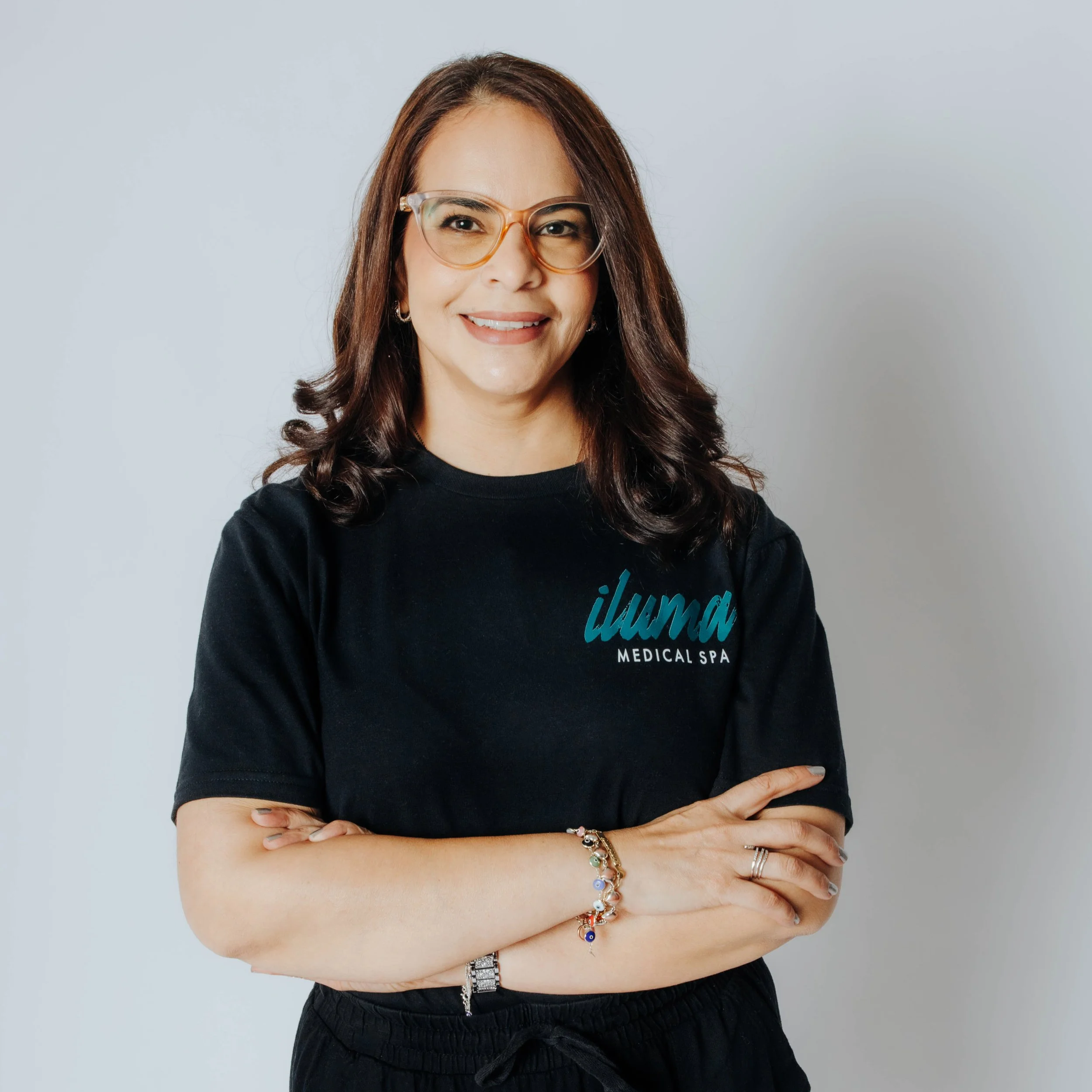 Staff member at ILUMA Medical Spa in Monrovia: a woman wearing glasses and a black shirt with the text 'iluma Medical Spa' on it, standing against a plain white background, with dark curly hair, smiling with arms crossed.