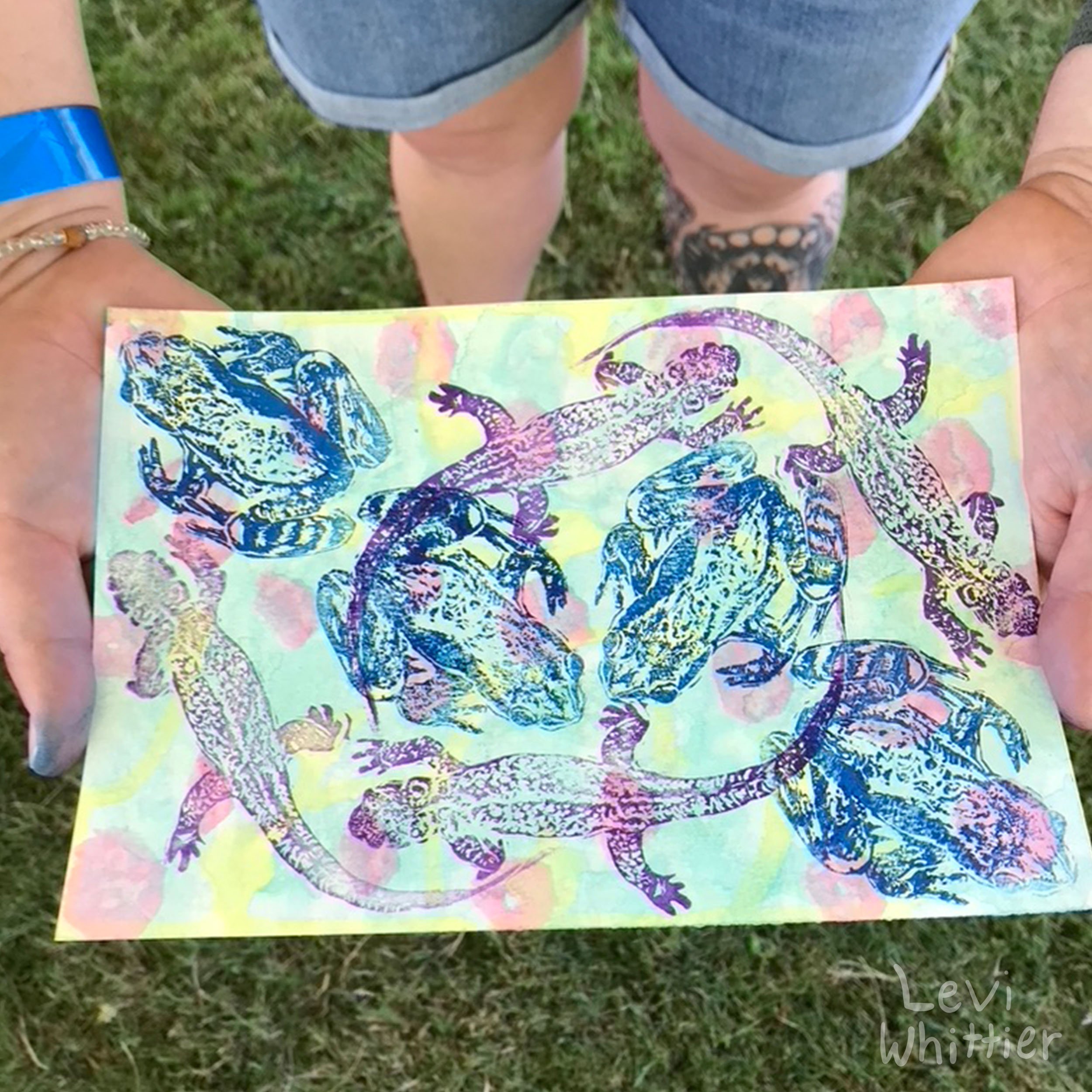 Colorful paper with multiple blue and purple lizard and frog prints, held by two hands in front of a grassy area.