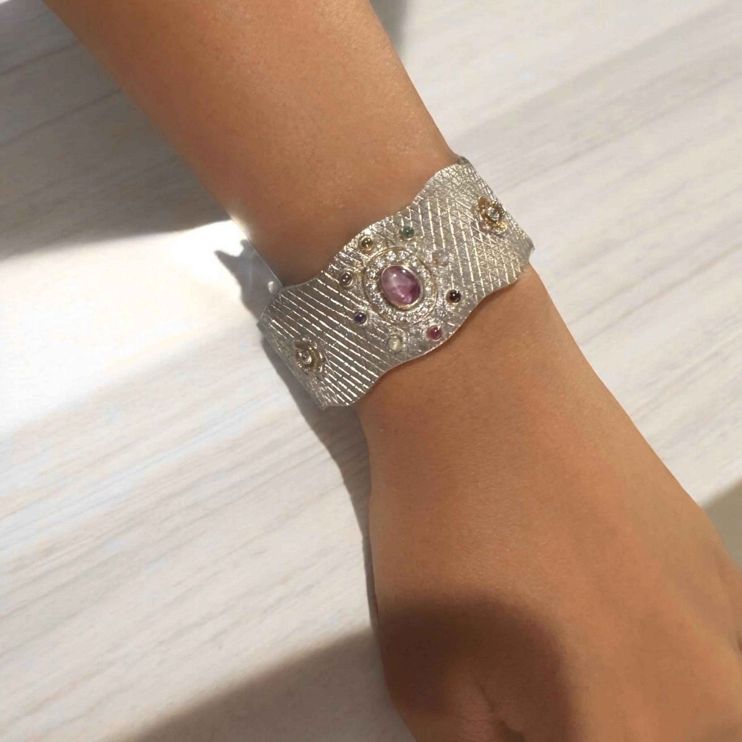 A person's wrist wearing a wide silver bracelet with a textured pattern and colorful gemstones.