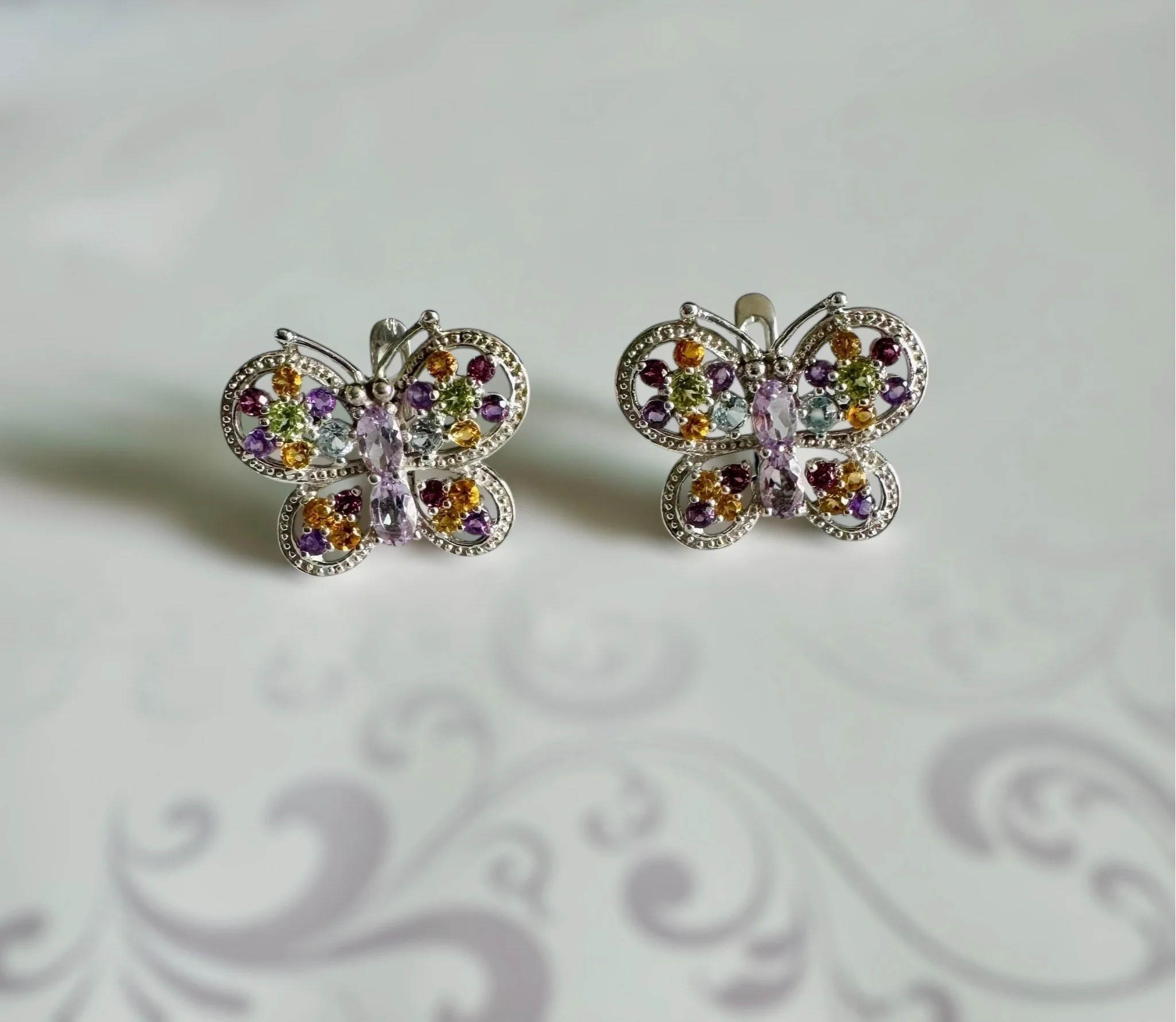 Silver butterfly earrings with multicolored gemstones on a light background with a decorative pattern.