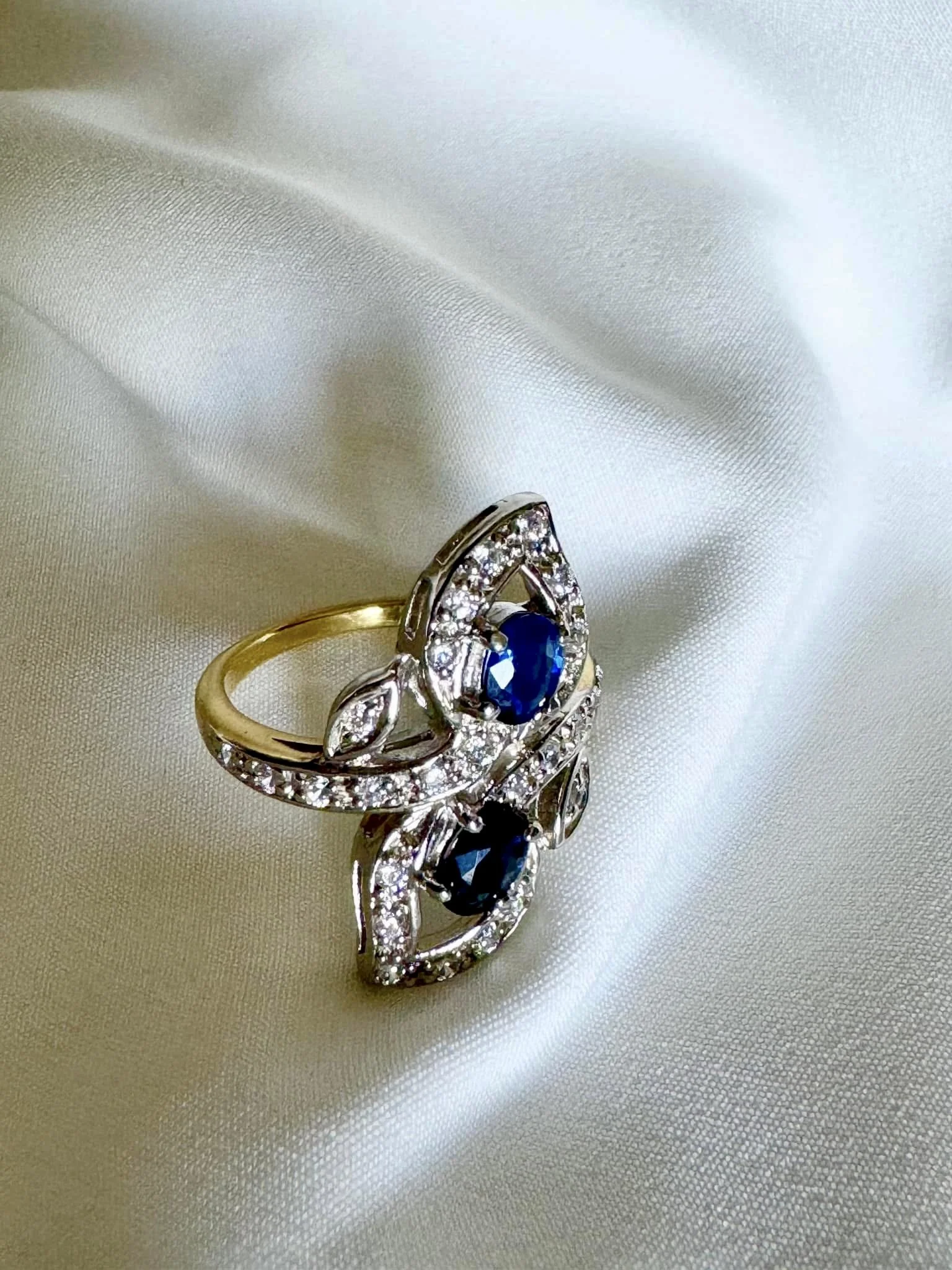 Engagement ring with two blue sapphires and many small diamonds set in silver and gold bands, resting on a silky white fabric.