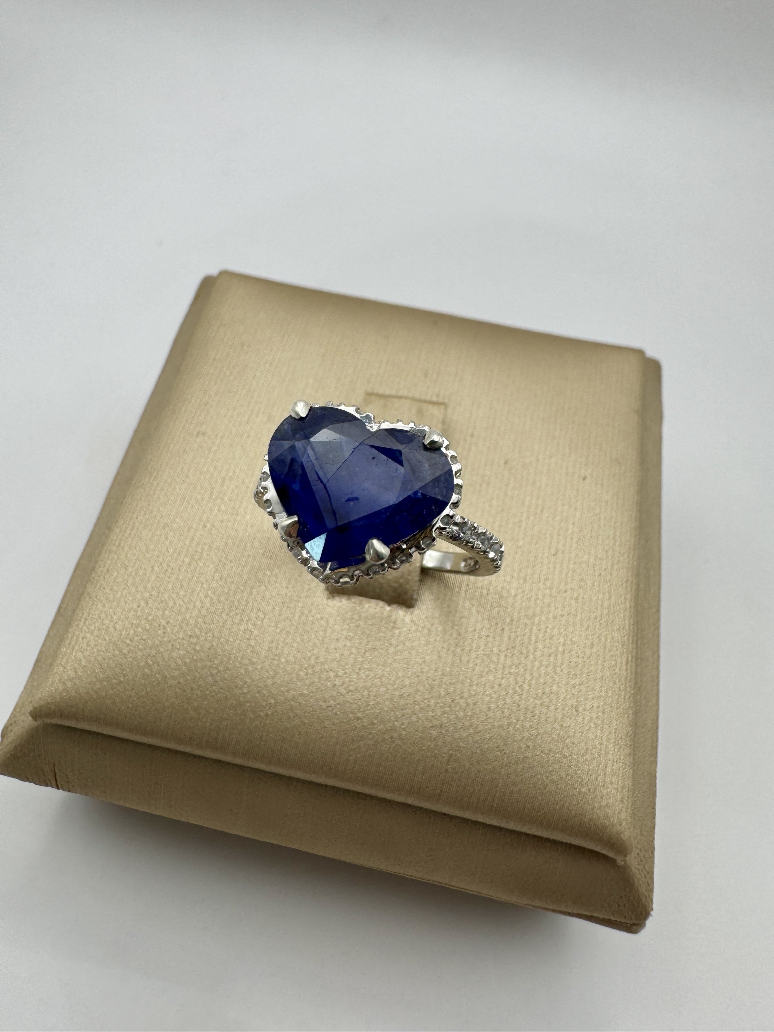 Silver ring with a large heart-shaped blue gemstone on a beige jewelry box.