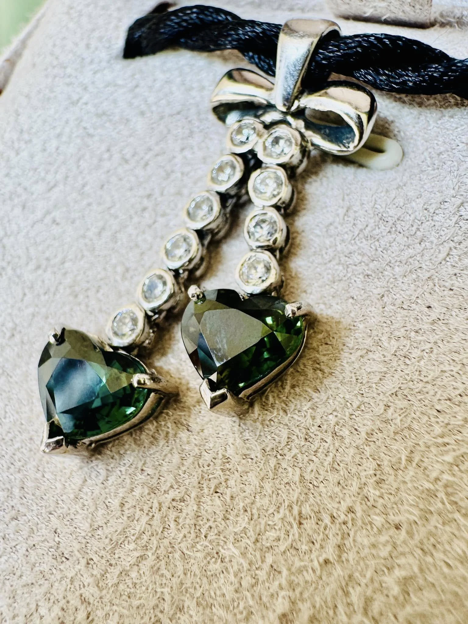 A heart-shaped green gemstone necklace with clear stones, attached to a silver chain with a black cord.