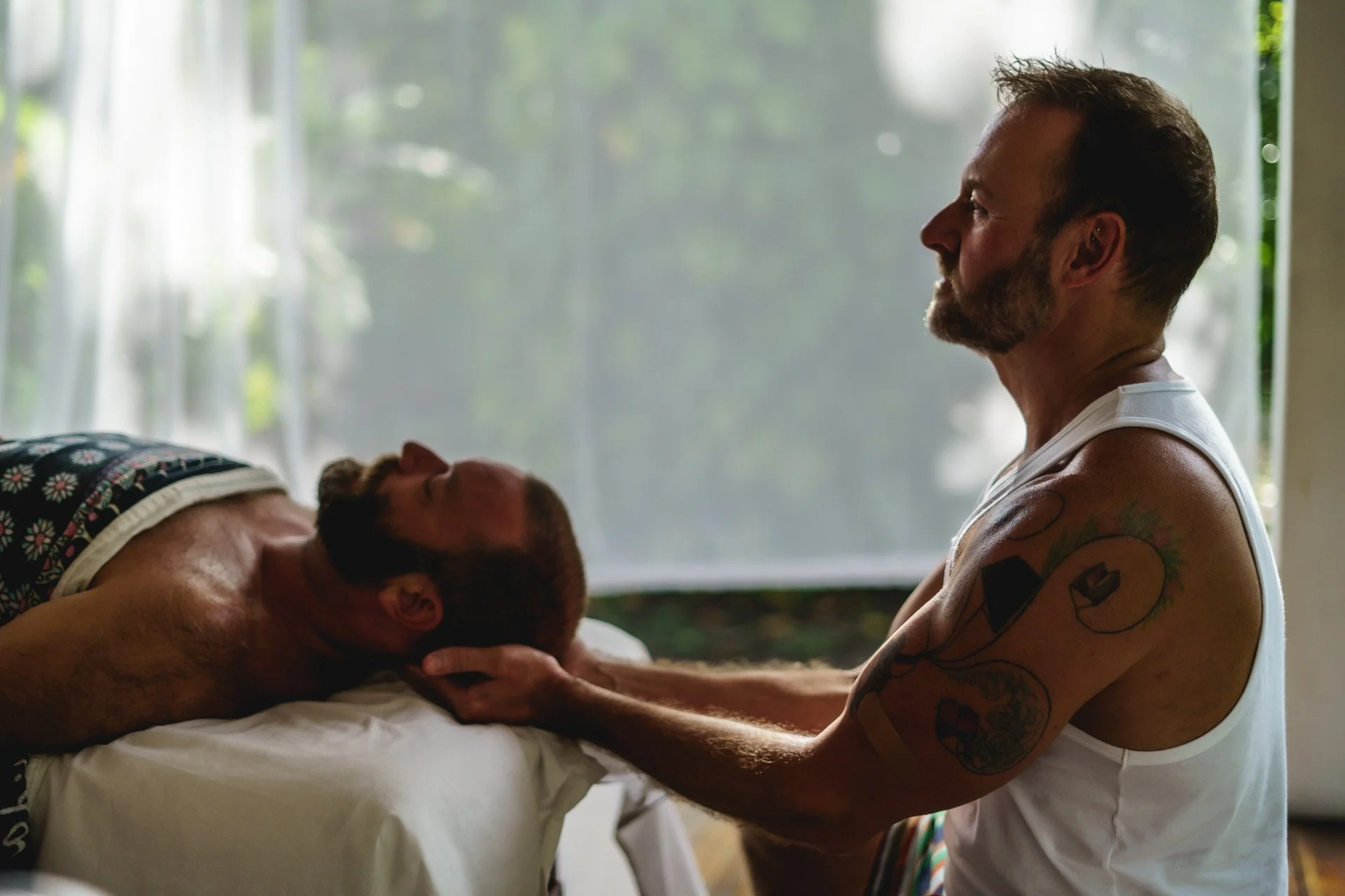 A man with tattoos is holding the head of another man lying on a bed, gently supporting his head while the man on the bed is relaxed with closed eyes, suggesting a moment of care or comfort. Encore Touch New Orleans Massage Therapy.