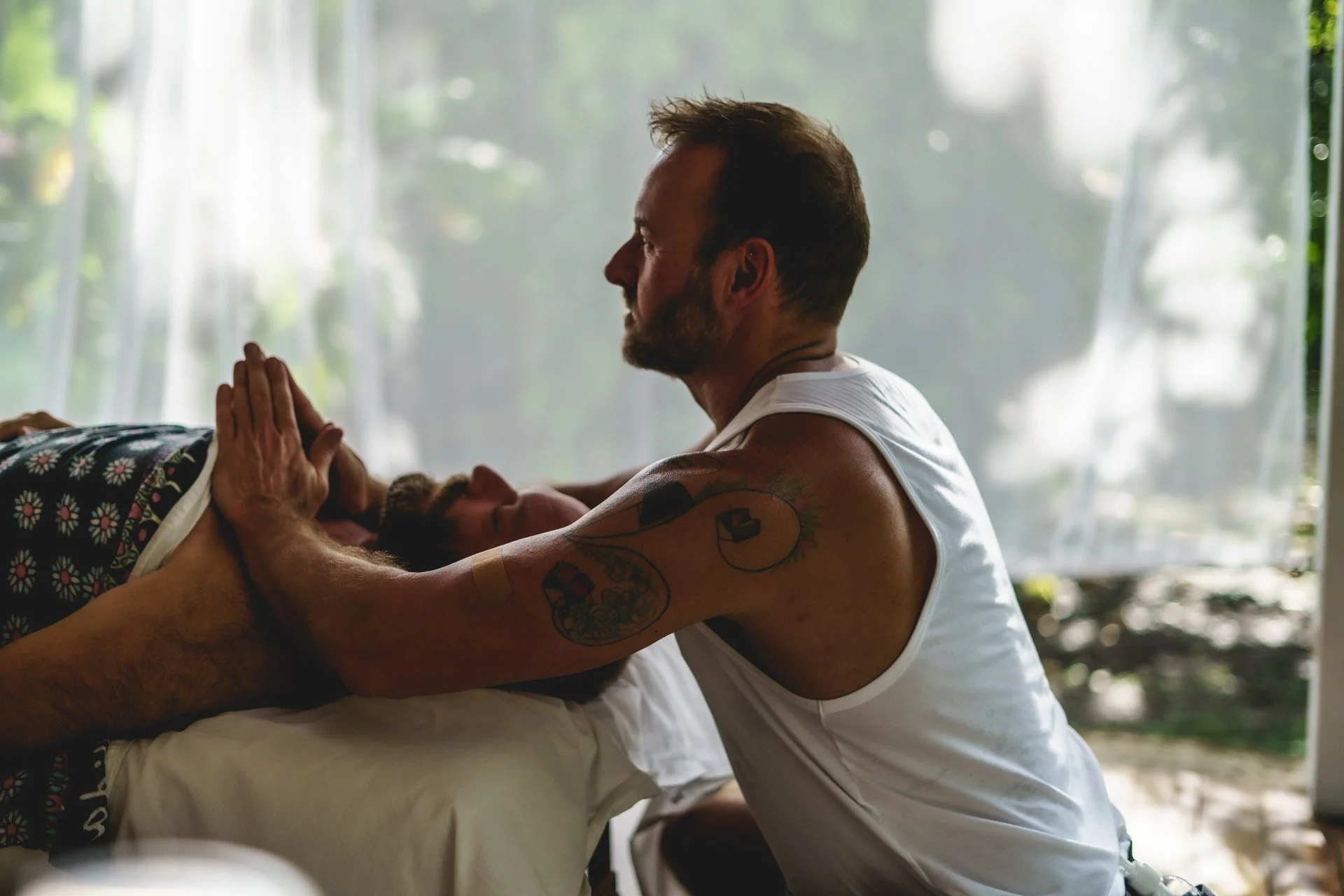 Man with tattoos in a white tank top performing a healing or reiki session on a person lying on a bed. Encore Touch New Orleans Massage Therapy.